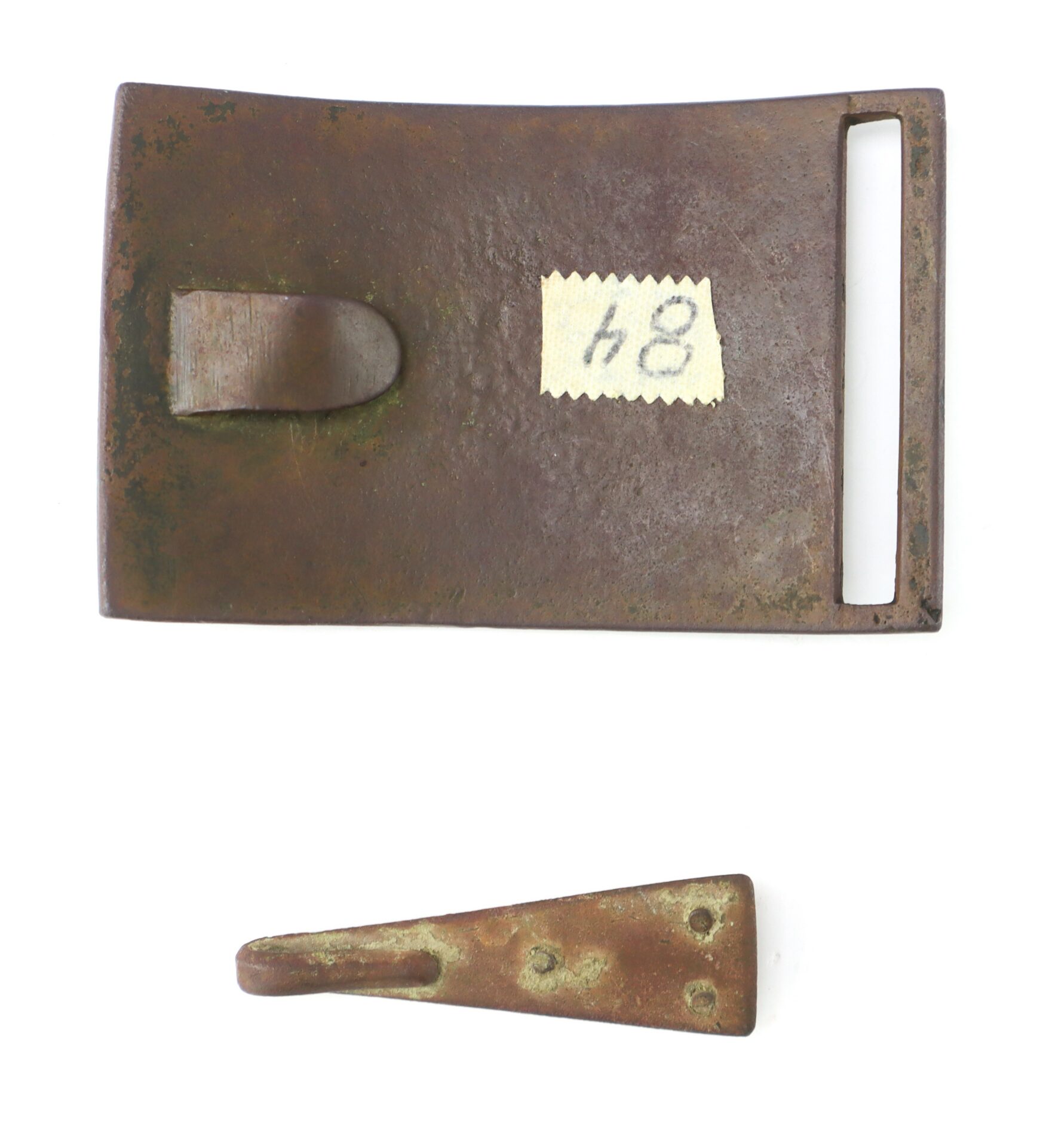 A rectangular bronze belt buckle with a rectangular slot and a detached, tapered tongue, both showing signs of age and corrosion. A small label with "84" is attached to the buckle.