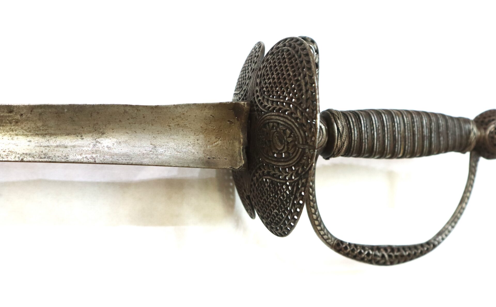 Close-up of an antique sword with an ornate, perforated metal guard and a ribbed handle, shown against a plain white background.