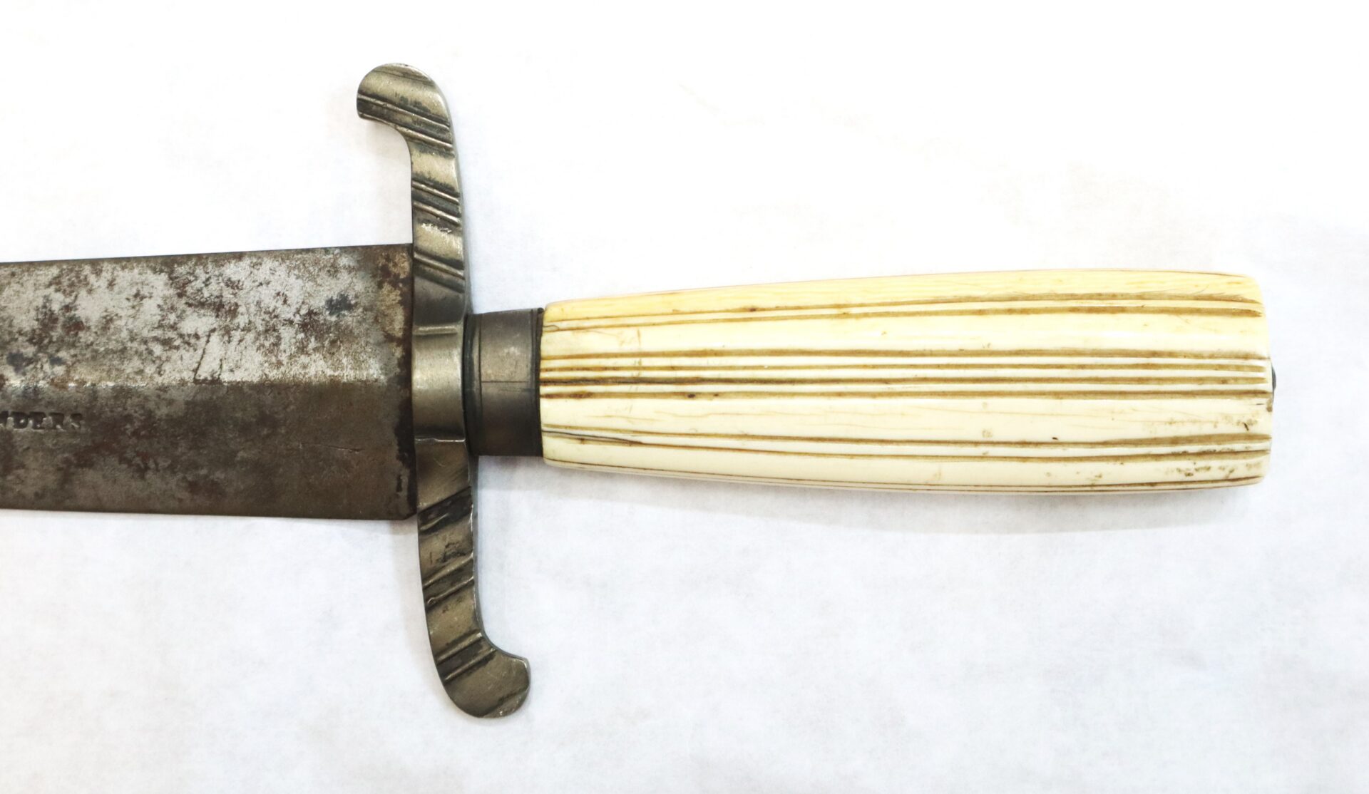 Close-up of an old knife with a tarnished metal blade, a simple cross guard, and a cream-colored, ribbed handle.