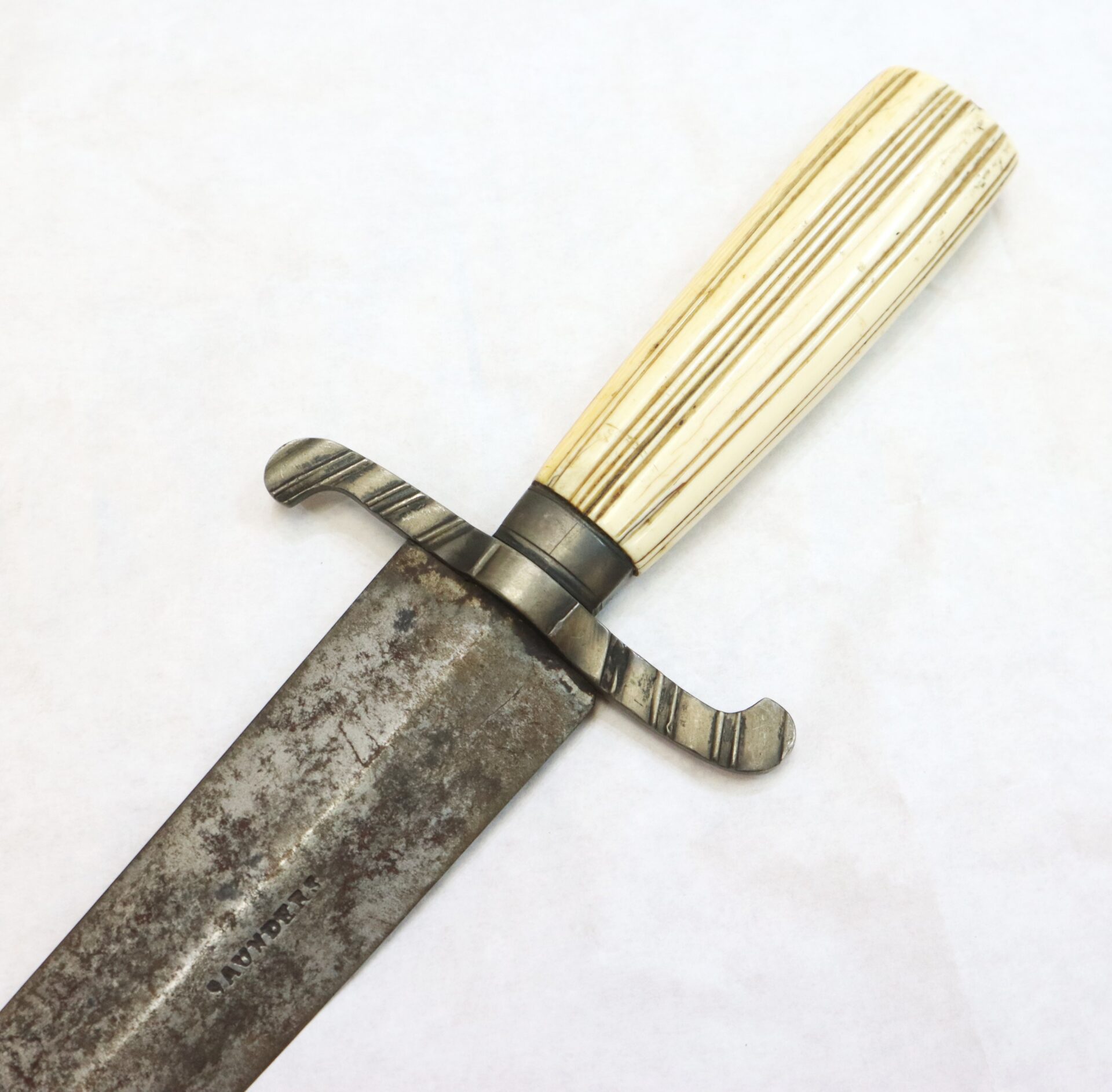 A close-up of a vintage knife with a stained, worn blade, a metal crossguard, and a ribbed, ivory-colored handle.
