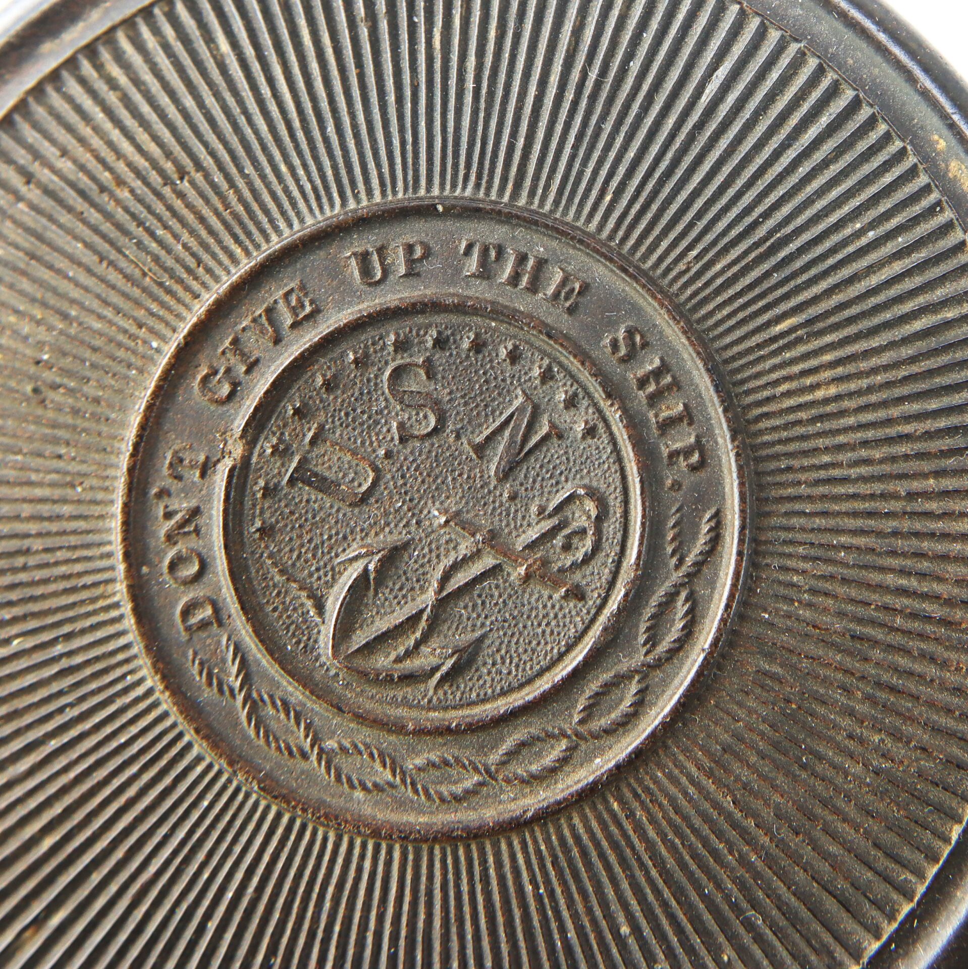 Close-up of a metal medallion engraved with "DON'T GIVE UP THE SHIP" and "U.S.N." above an anchor, surrounded by textured and radiating lines.
