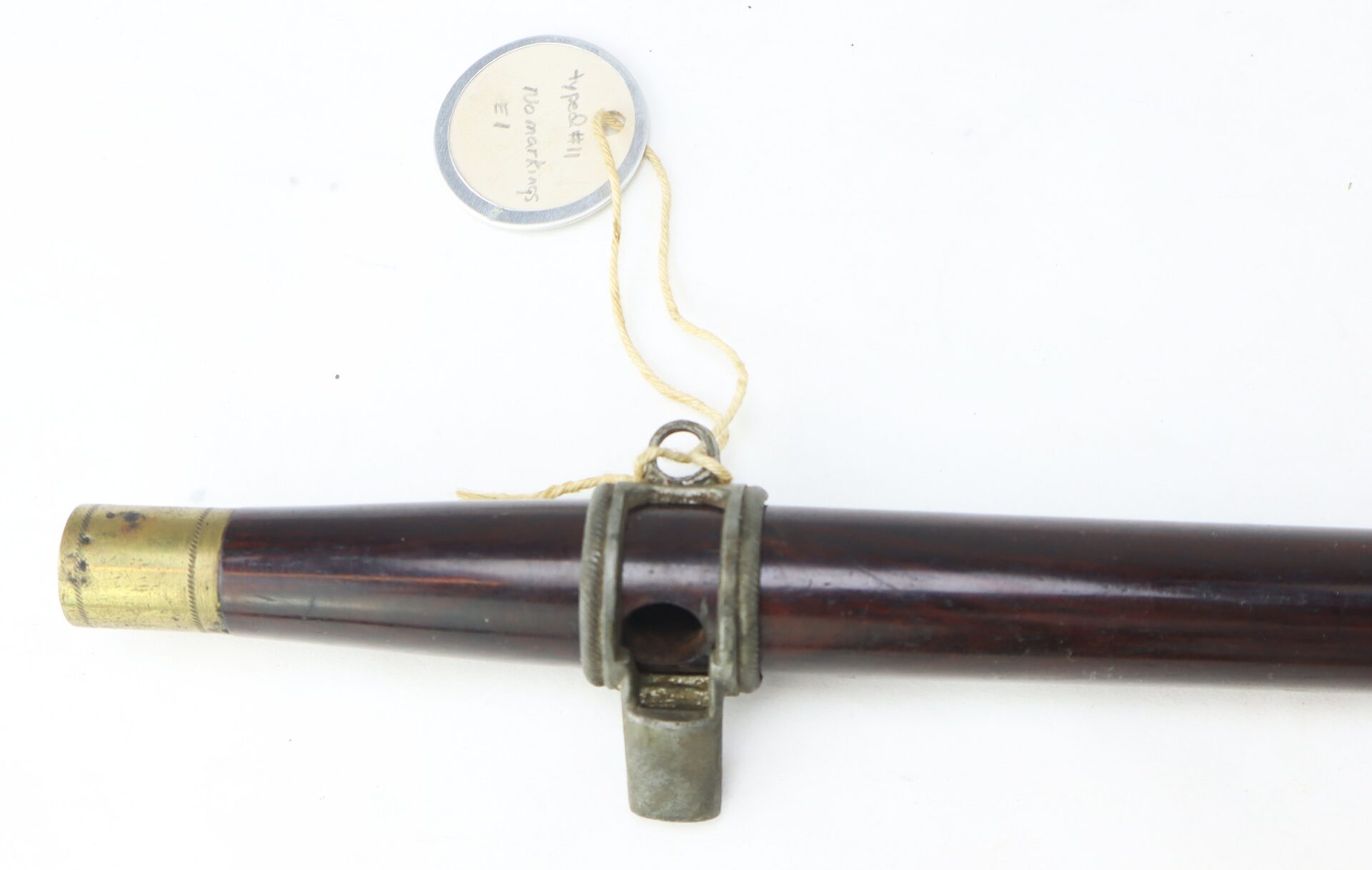A close-up of a Civil War–Era Fife with Brass Fittings, Gettysburg Provenance, featuring a metal fitting and a circular tag with handwritten text attached to a string.