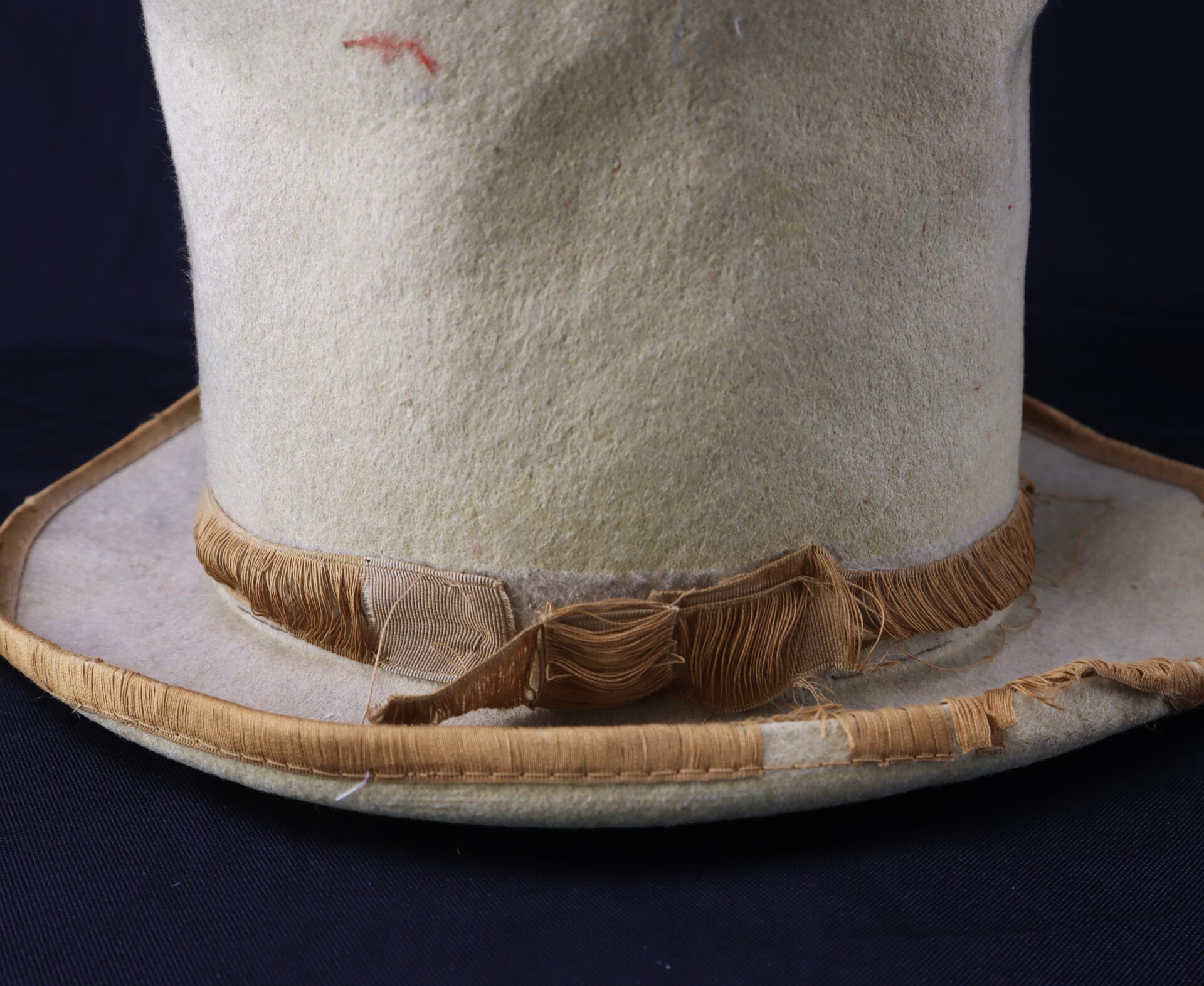 A close-up of the Awesome Top Hat for the 1888 Republican Campaign, featuring a light color and frayed brown ribbon, resting on a dark surface.