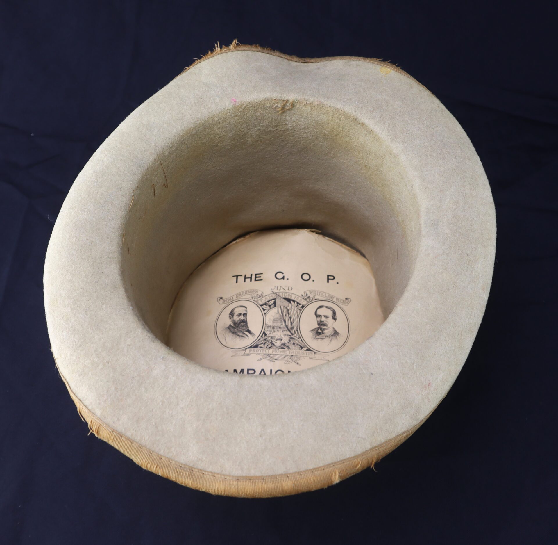 The "Awesome Top Hat for the 1888 Republican Campaign" holds a campaign flyer featuring two men's portraits labeled "THE G.O.P." and partial text, resting inside the upturned hat on a dark surface.