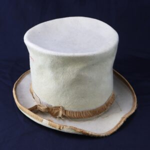 An off-white Awesome Top Hat for the 1888 Republican Campaign features a frayed gold ribbon and a misshapen brim, set against a dark background.