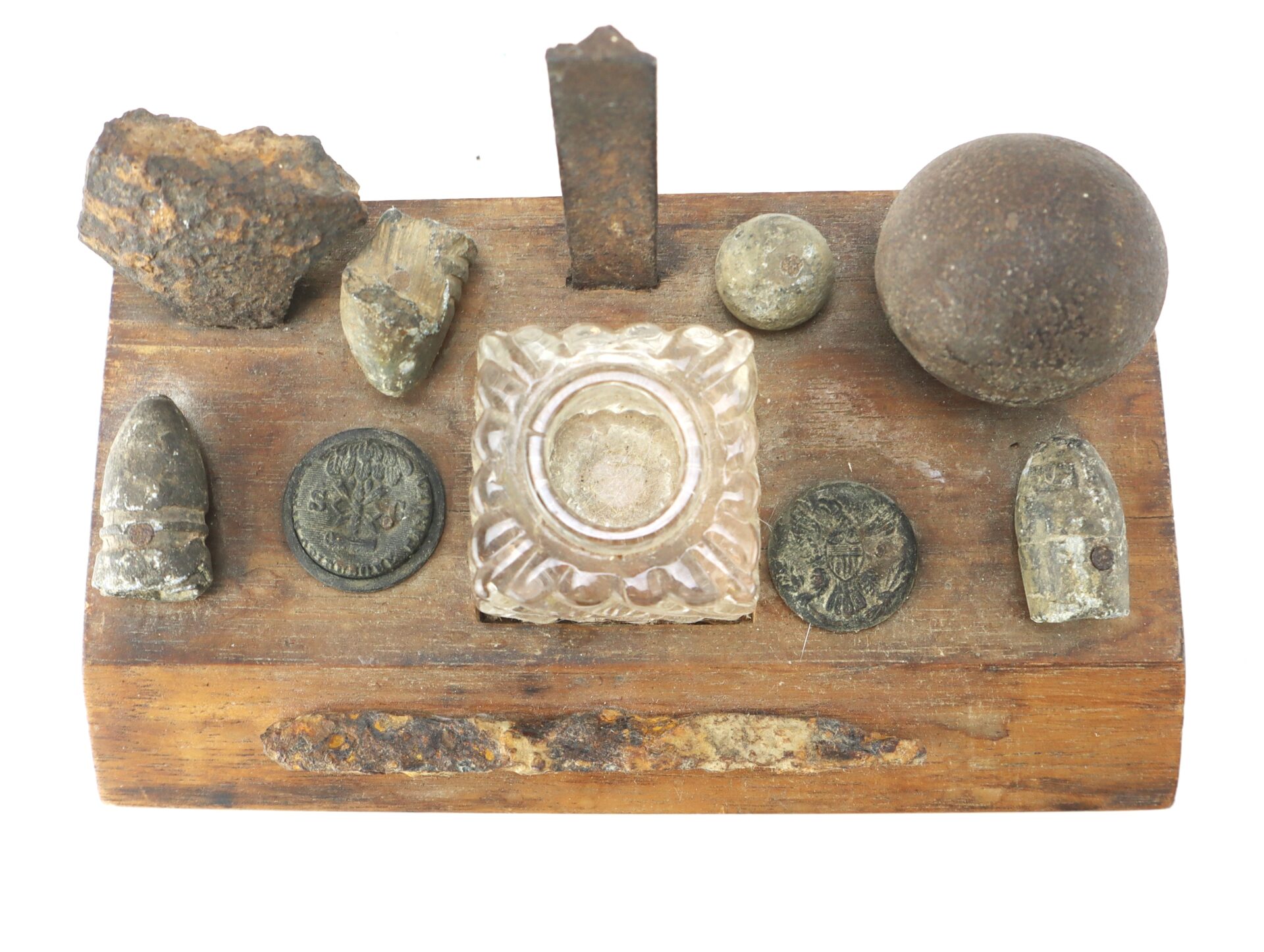A wooden display holds old bullets, coins, metal fragments, a glass inkwell, and a metal ball, arranged in rows on a white background.