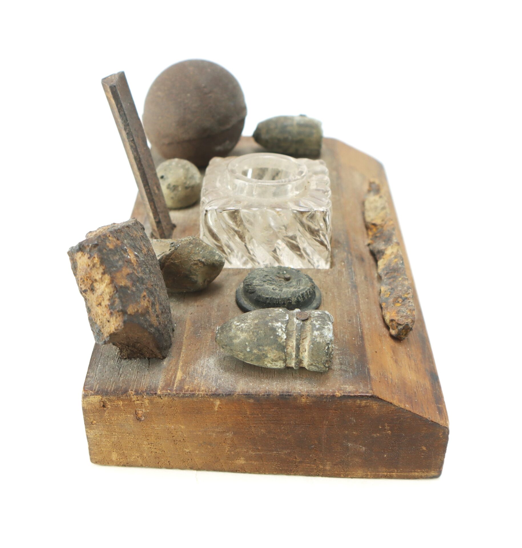A wooden display holds various old metal objects, a glass inkwell, lead bullets, a round ball, and a rusted metal rod arranged on top.