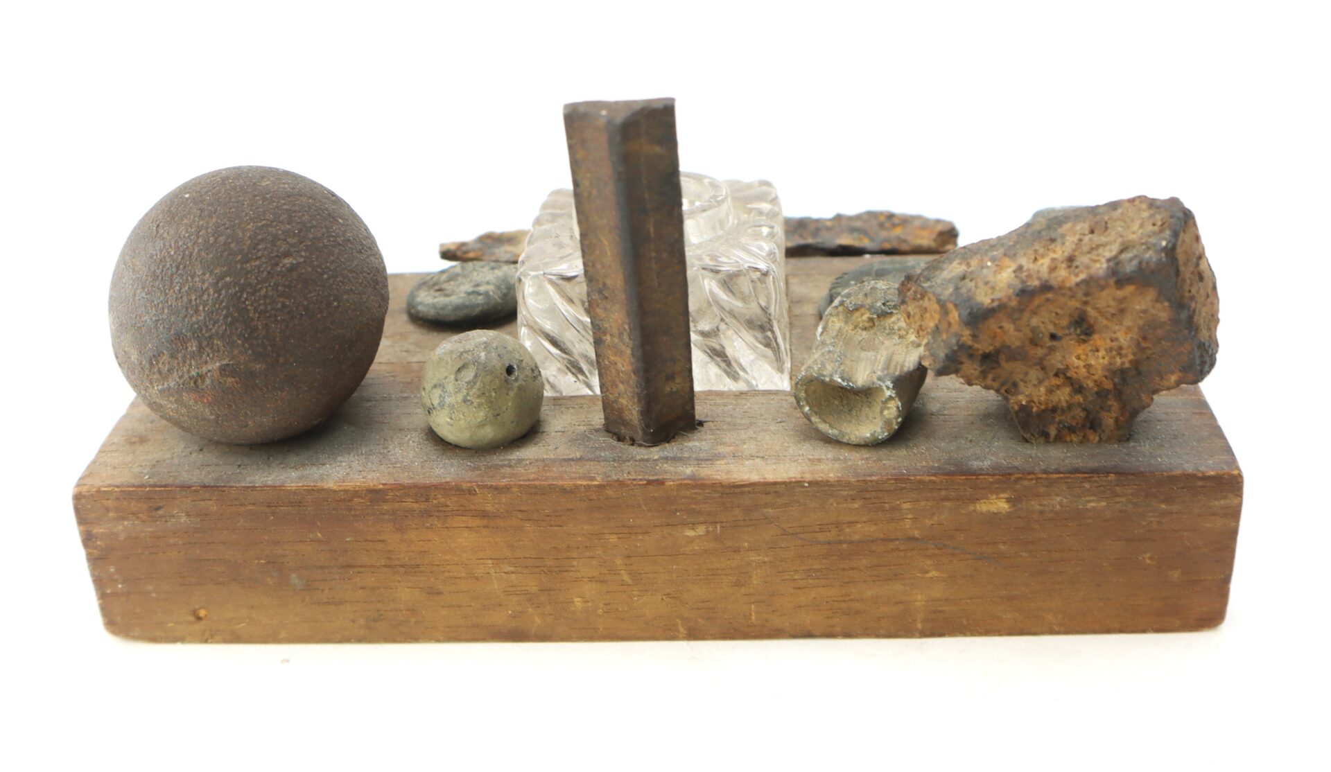 A wooden base displaying various objects, including a metal sphere, a glass block, a rusted metal piece, and several small round and irregular metal fragments.