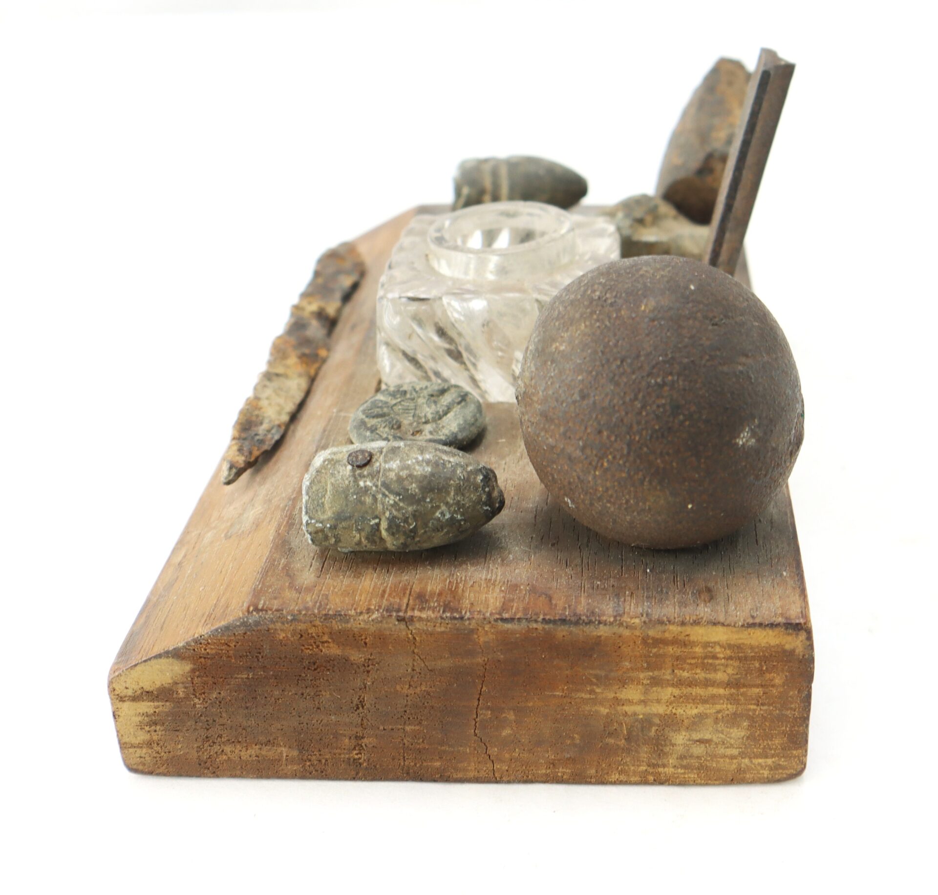A wooden board displays assorted old metal objects, including bullets, a glass inkwell, a nail, and a rusted iron spike, all arranged in a row on a white background.
