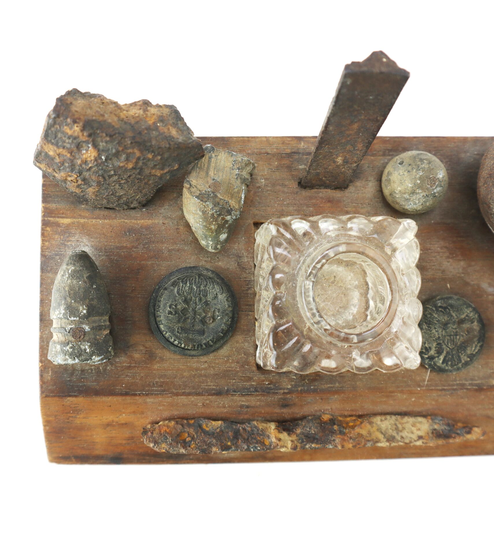 A wooden board displays old coins, bullets, rocks, a glass inkwell, and a rusty metal object arranged in a row.
