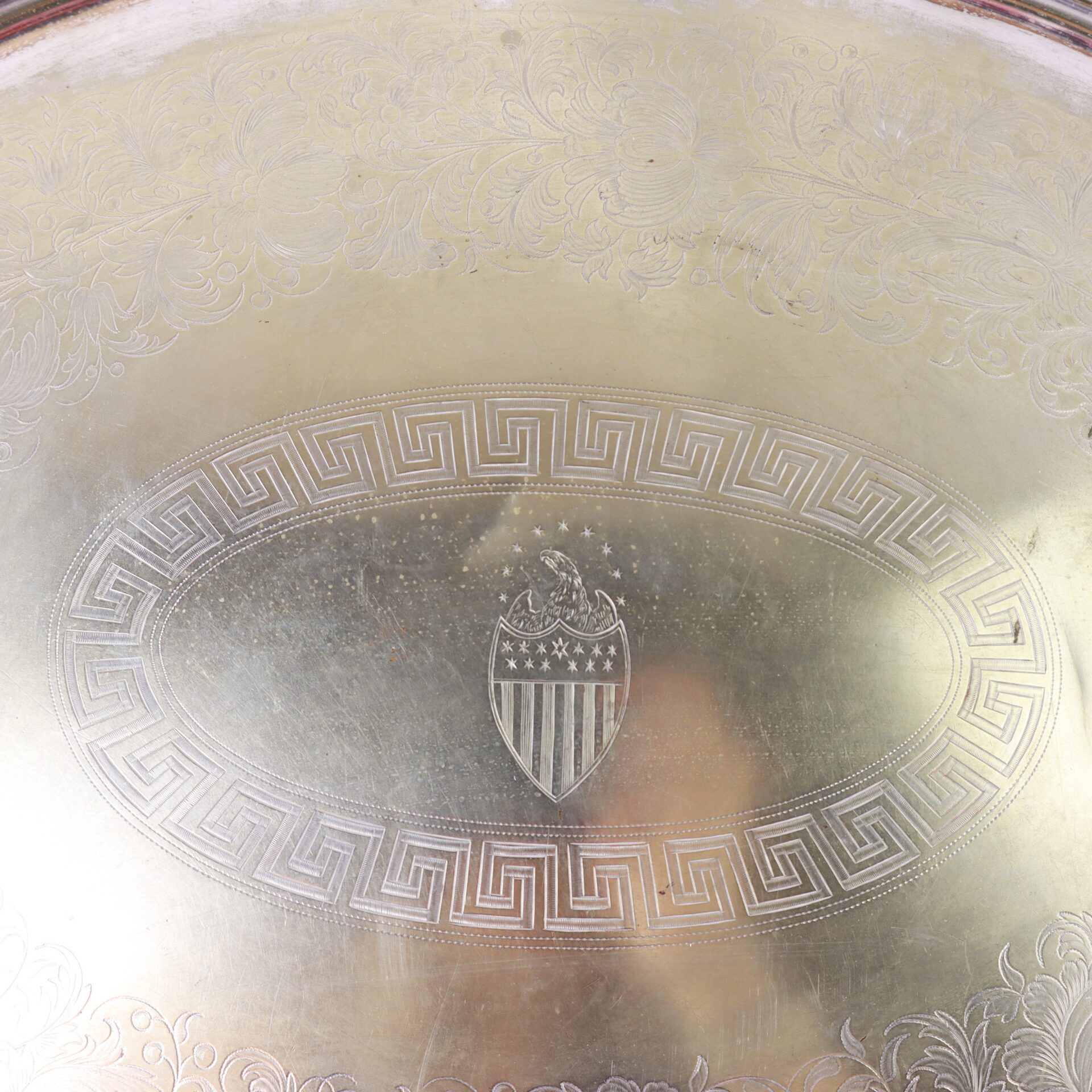 A metal tray with an engraved border, featuring a central oval containing a shield emblem with stars, stripes, and an eagle.