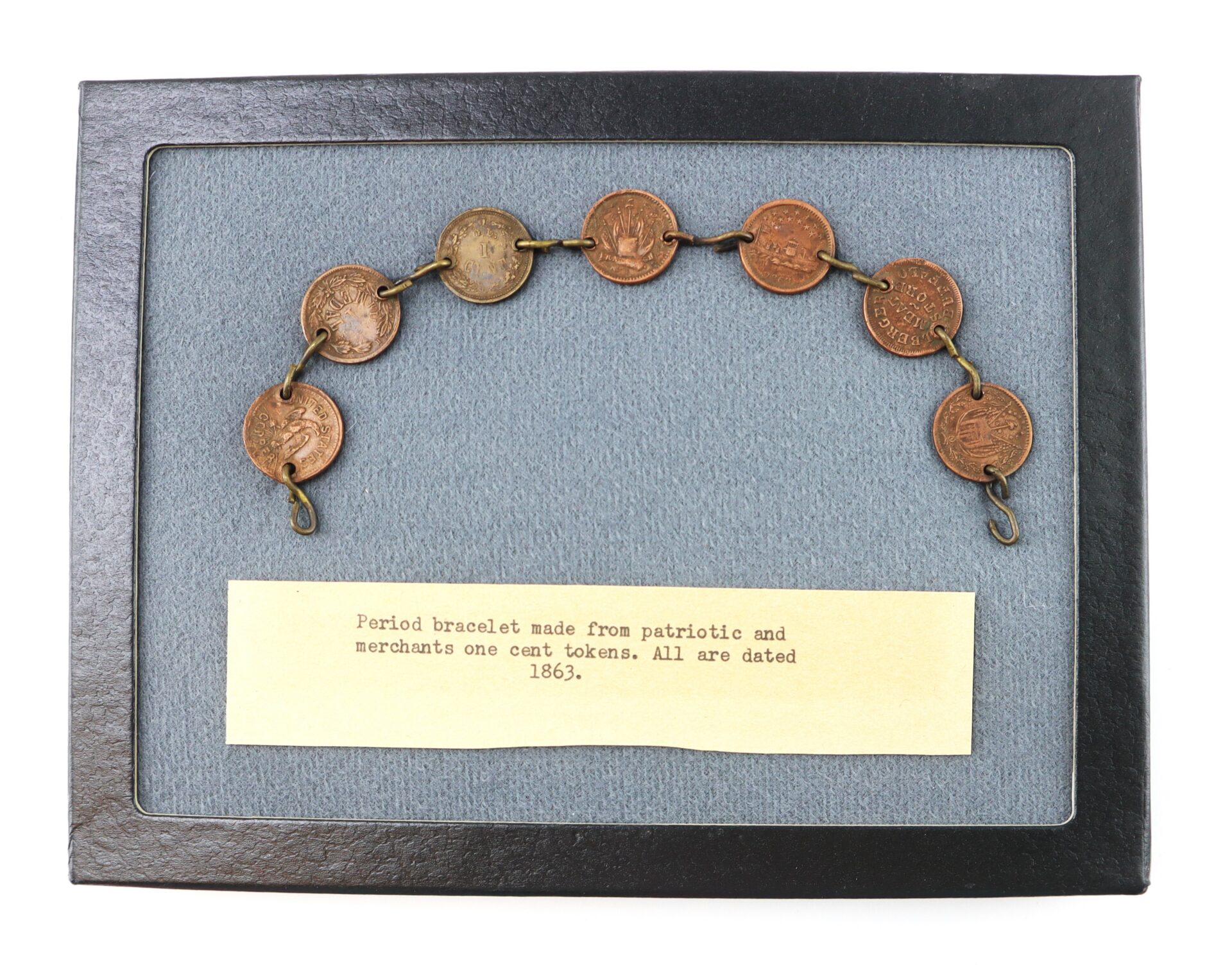 A bracelet made from seven linked one cent tokens displayed in a frame with a label reading, “Period bracelet made from patriotic and merchants one cent tokens. All are dated 1863.”.