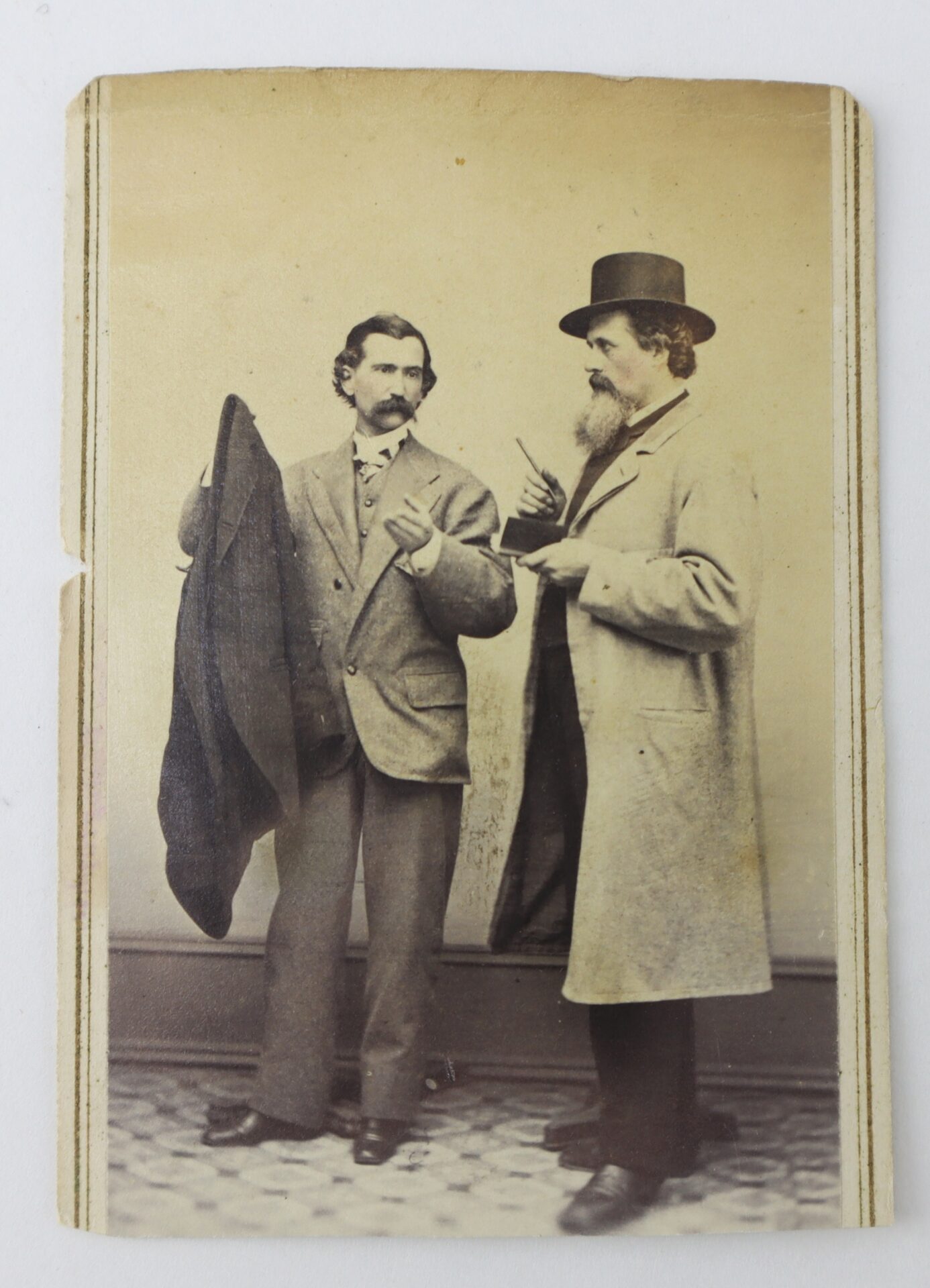 Two men in 19th-century attire stand indoors; one holds a coat, the other, wearing a top hat, points with a cane or stick.