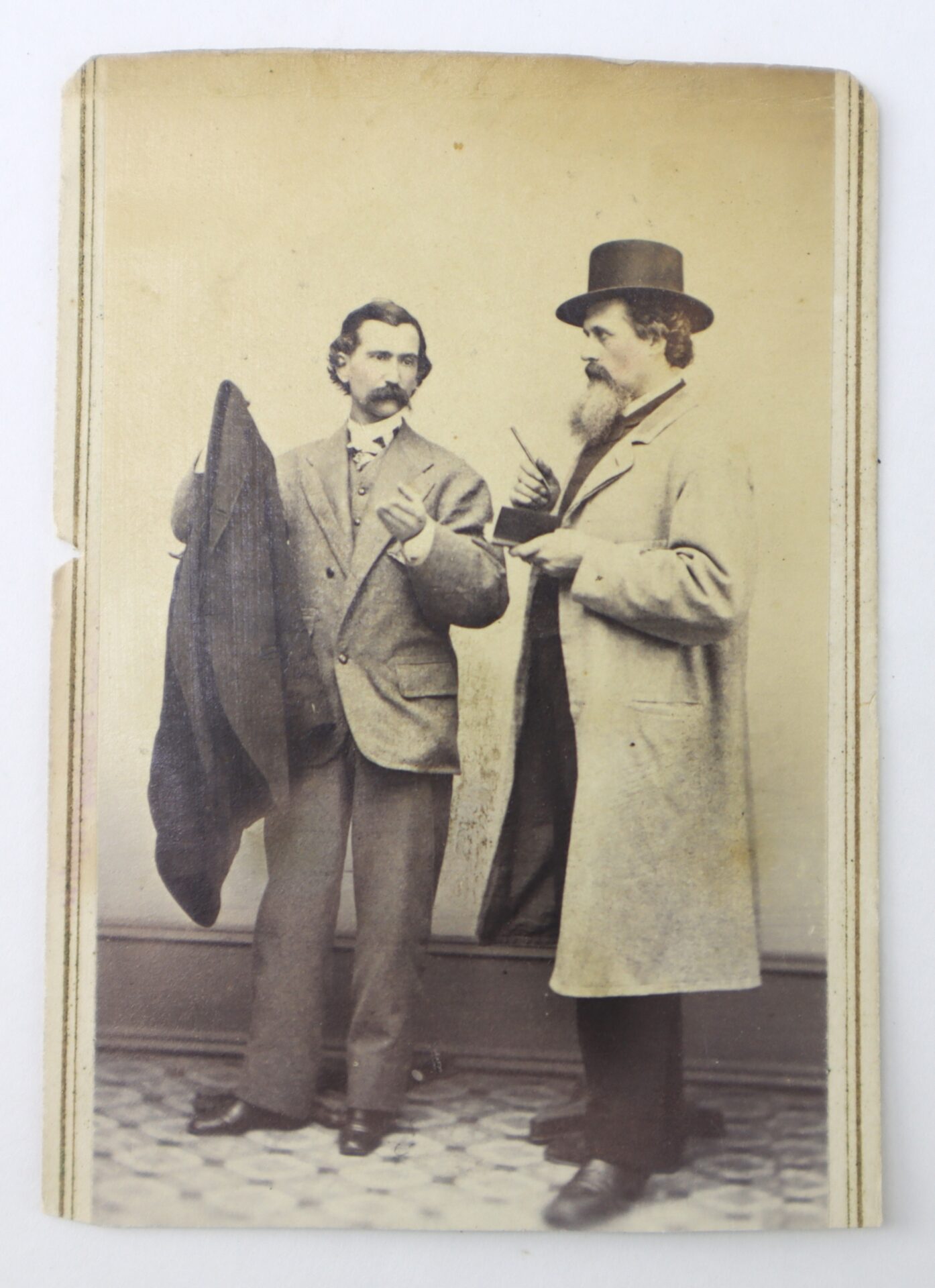 Two men in 19th-century attire stand indoors, one holding a coat and the other wearing a top hat and long coat, engaged in conversation.