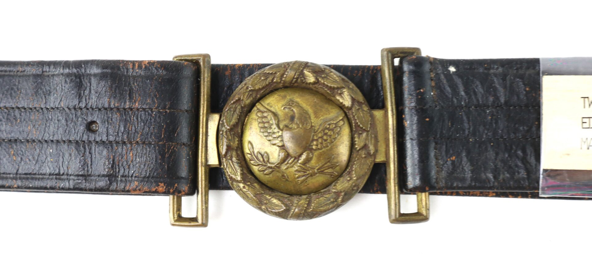 A worn black leather belt with a brass oval buckle featuring an embossed eagle and decorative leaves.