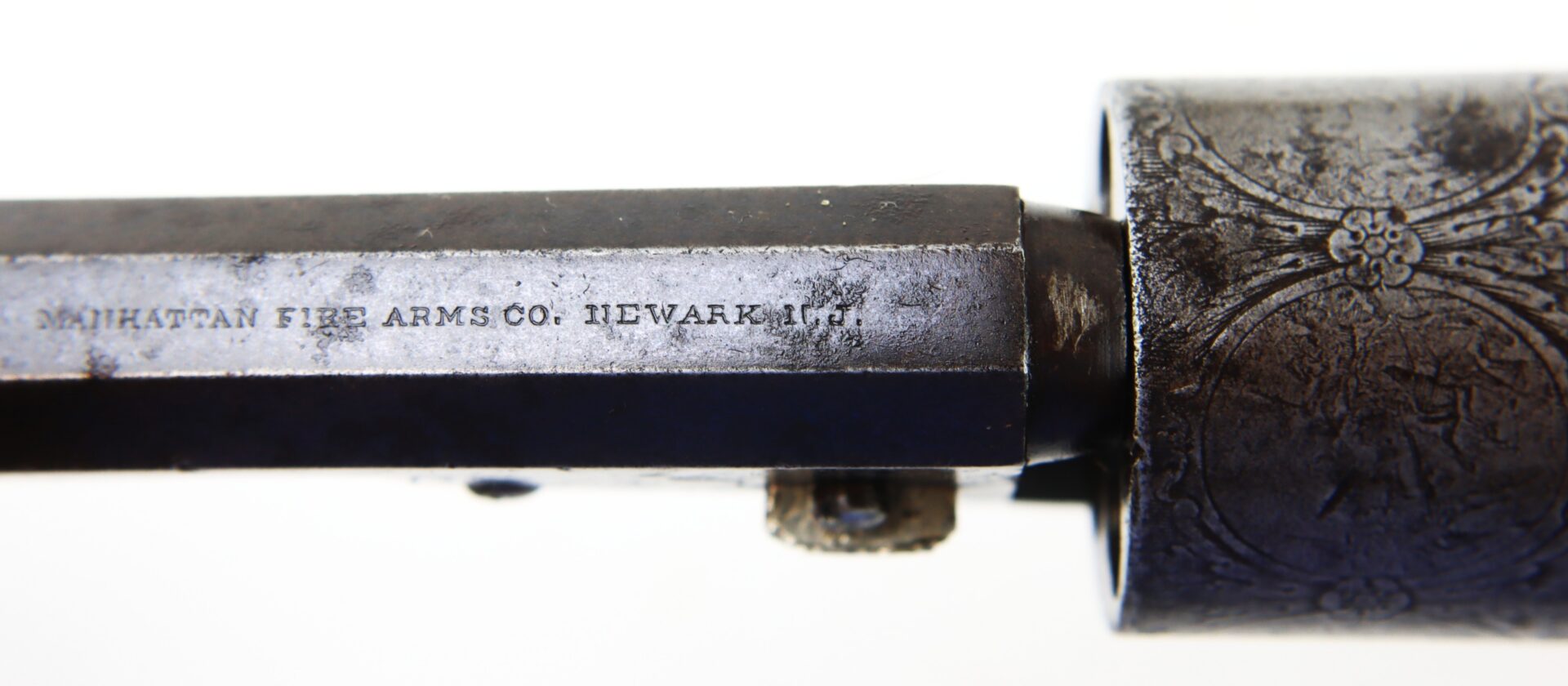 Close-up of a metal firearm barrel engraved with "Manhattan Fire Arms Co. Newark N.J." and part of an engraved cylinder.