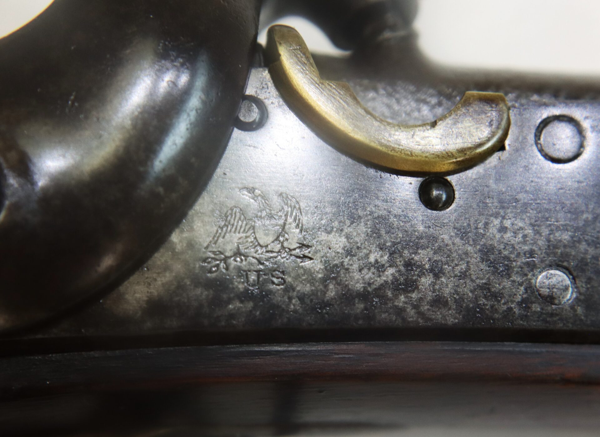 Close-up of a metal firearm with an engraved eagle, the letters “U.S.” beneath it, and part of a brass trigger guard visible.