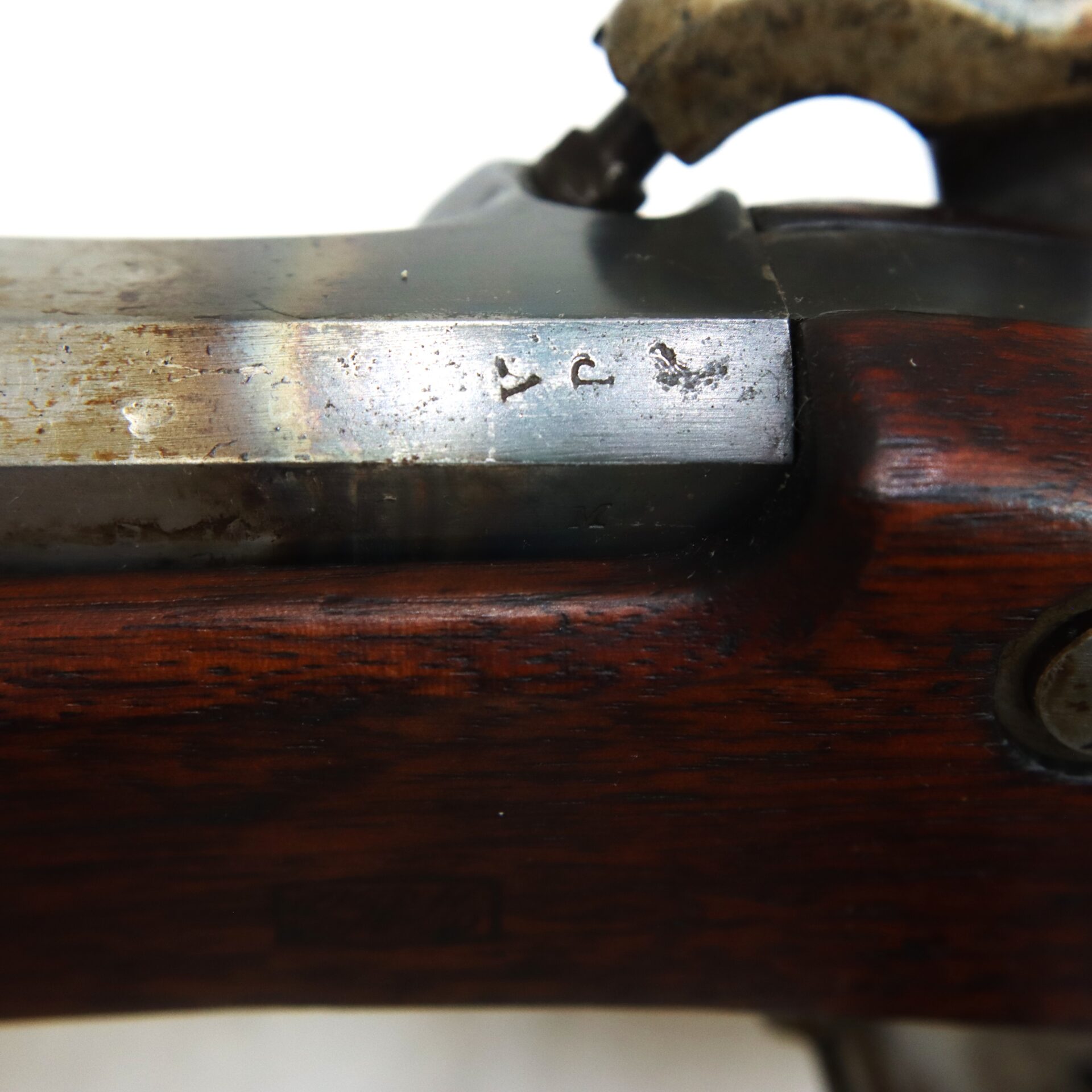 Close-up of a section of a rifle, showing part of the metal barrel with two small stamped marks and dark wood stock beneath.