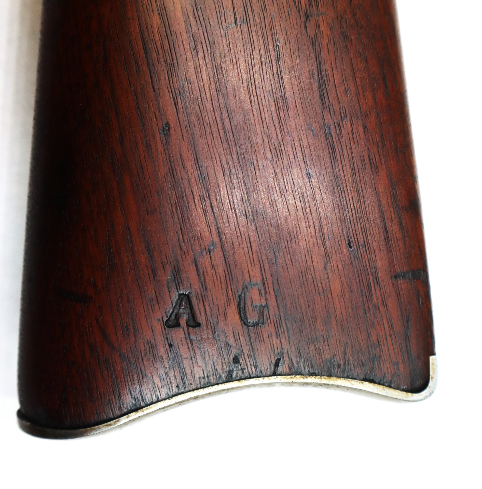 Close-up of a wooden rifle buttstock with the letters "AG" stamped near the metal buttplate.