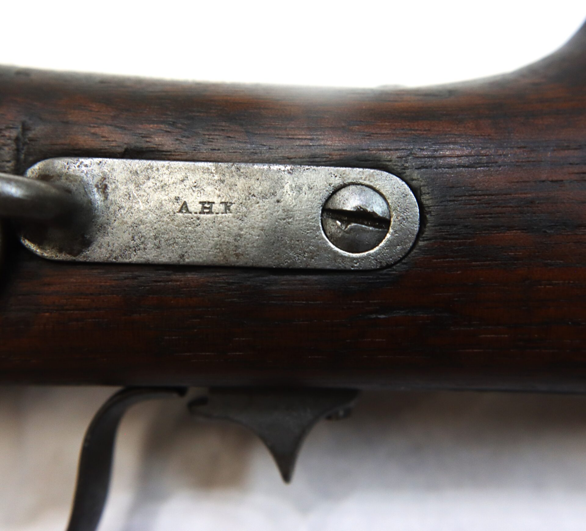 Close-up of a metal plate on a wooden rifle stock, engraved with the initials "A.H.K" and secured by a single flathead screw.