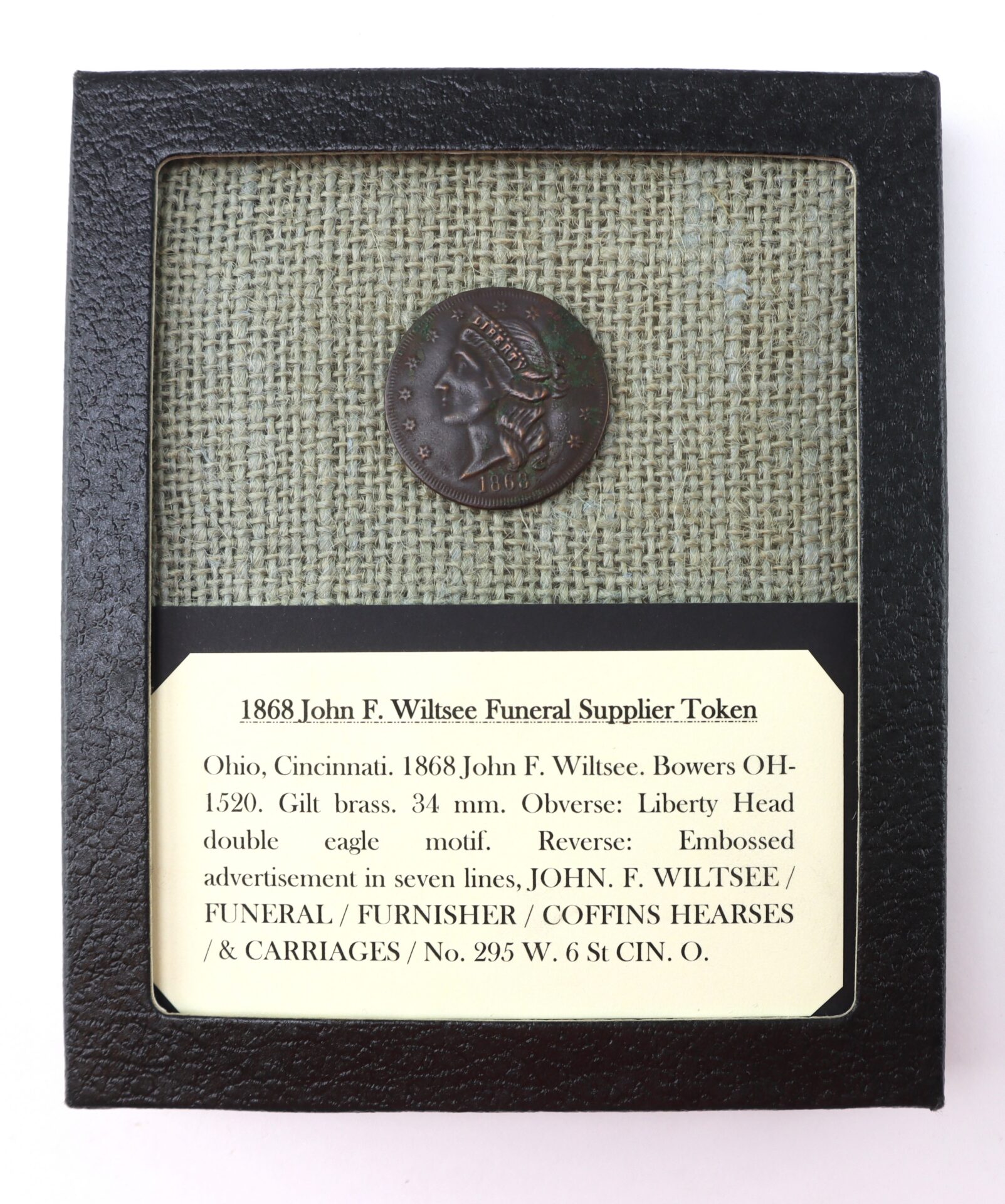 A framed 1868 John F. Wiltsee Funeral Supplier Token displayed with a label describing its origin, design, and advertising details.