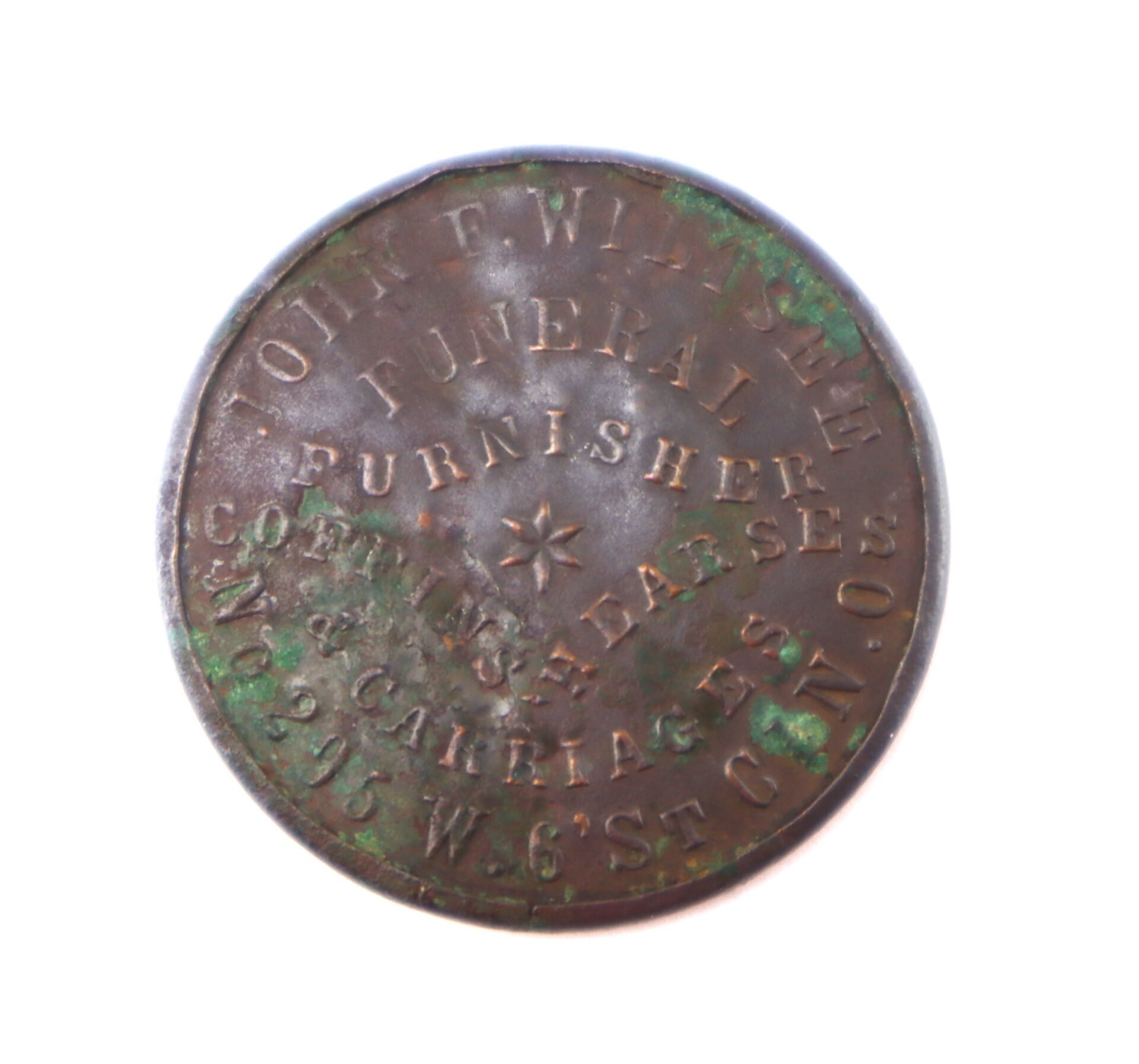 Round metal token inscribed with "John F. Winstone, Funeral Furnisher, Coffins, Hearses, and Carriages, 296 W. 6th St." Some tarnish and green corrosion visible.