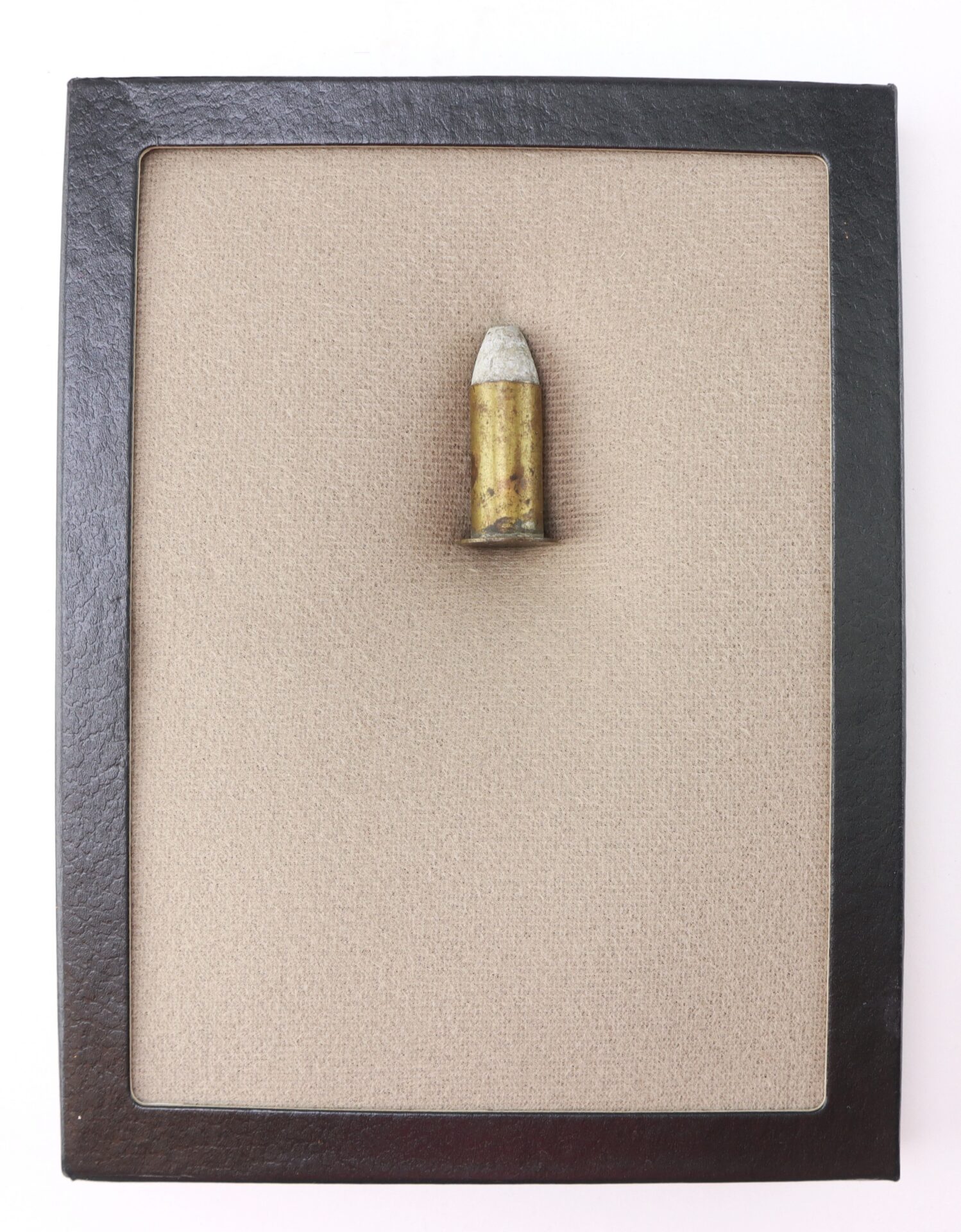 A single bullet fragment mounted upright on beige fabric, displayed in a rectangular black frame.
