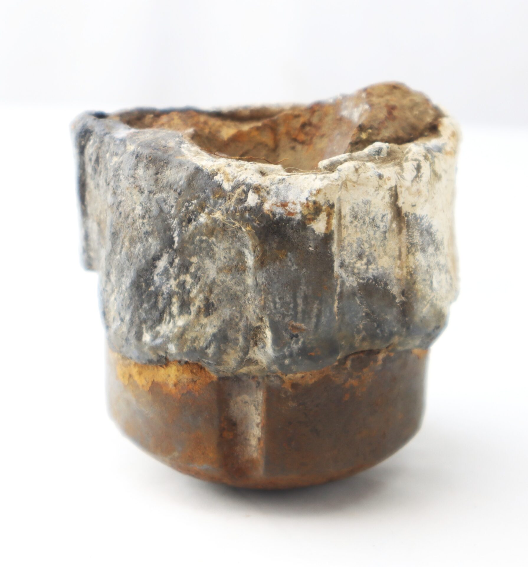 A corroded and partially melted metal object with a rough, uneven surface and a mix of rust, silver, and brownish colors.