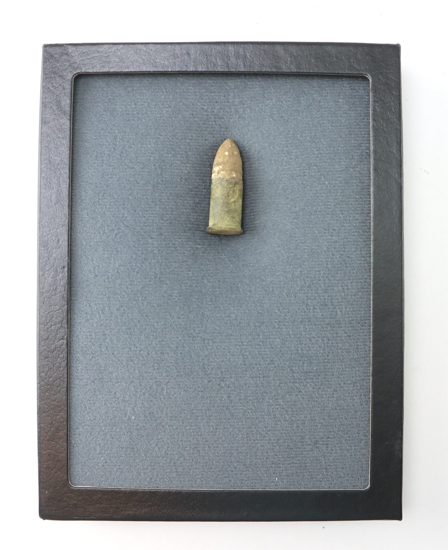 A single old bullet is displayed upright in the center of a black-framed shadow box with a textured gray background.