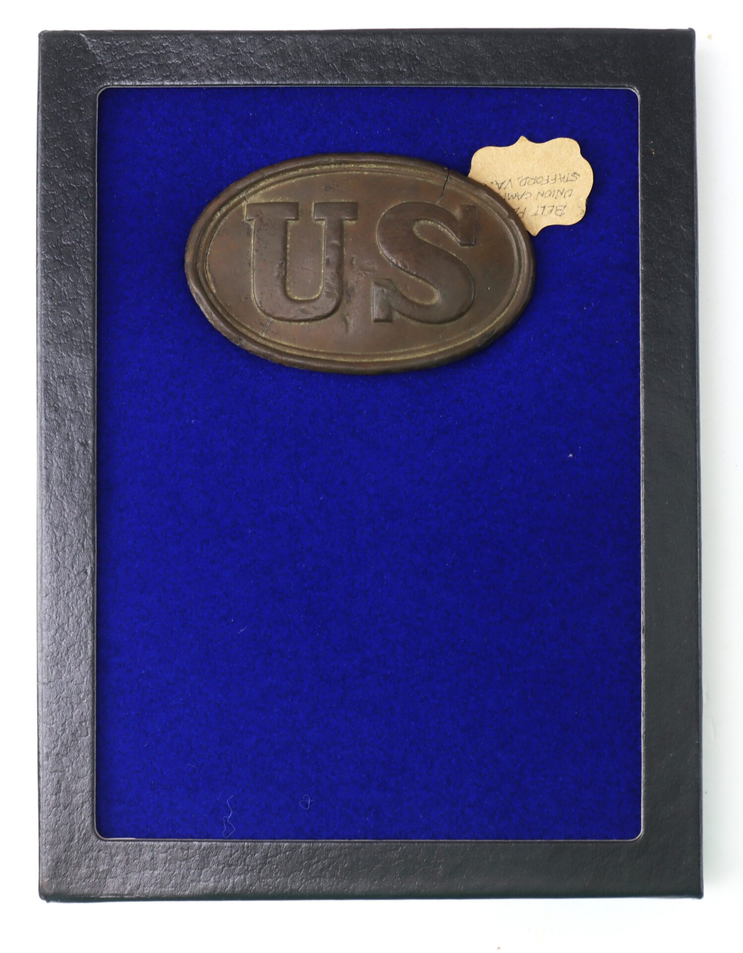 A rectangular display case with a blue background holds an oval brass "US" military belt buckle and a small paper label with handwritten text in the top right corner.