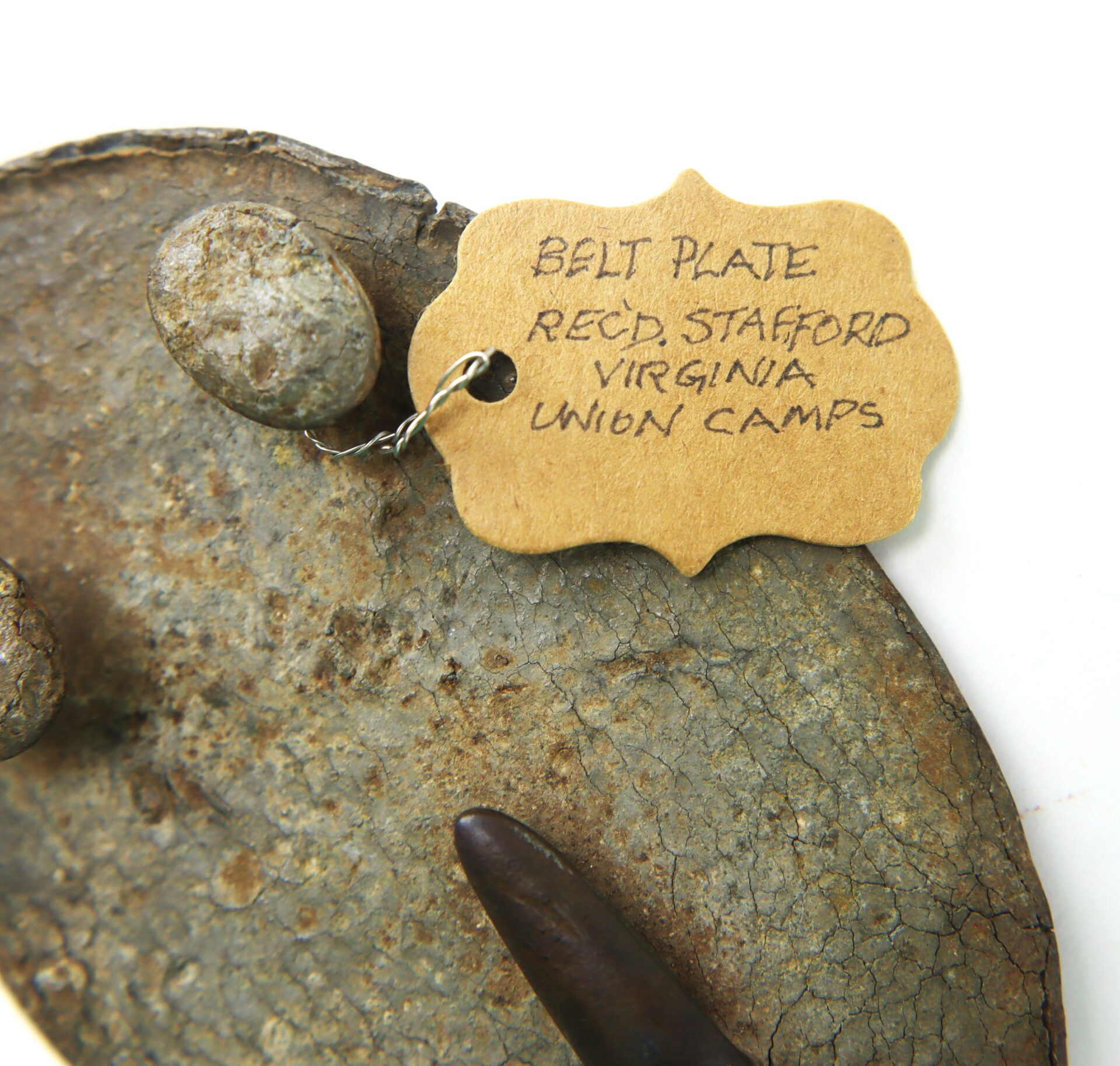 A metal artifact with a tag reading "Belt plate, Recd, Stafford, Virginia, Union camps" attached by wire.