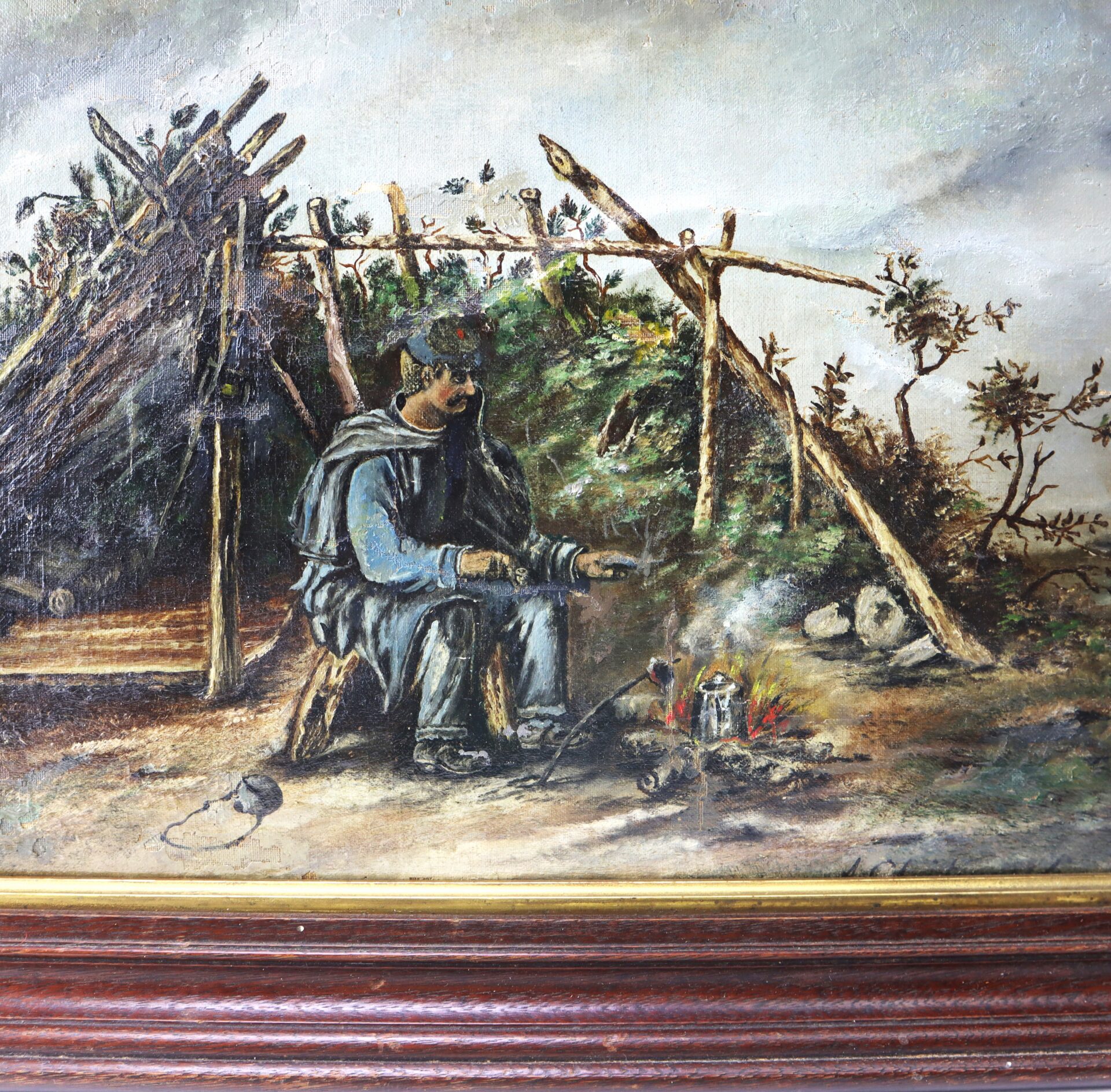 A man in rugged clothing sits by a small campfire with a kettle, next to a rustic shelter made of sticks and branches, surrounded by sparse vegetation.