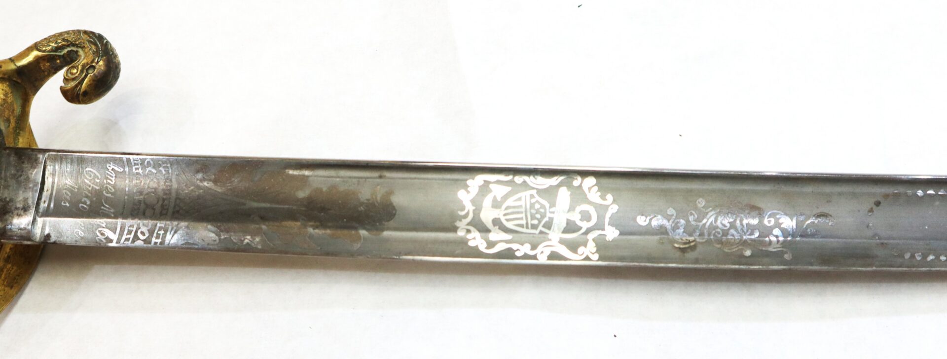 Close-up of a sword blade with engraved decorative patterns and designs, showing part of a gold-colored ornate guard on the left side.