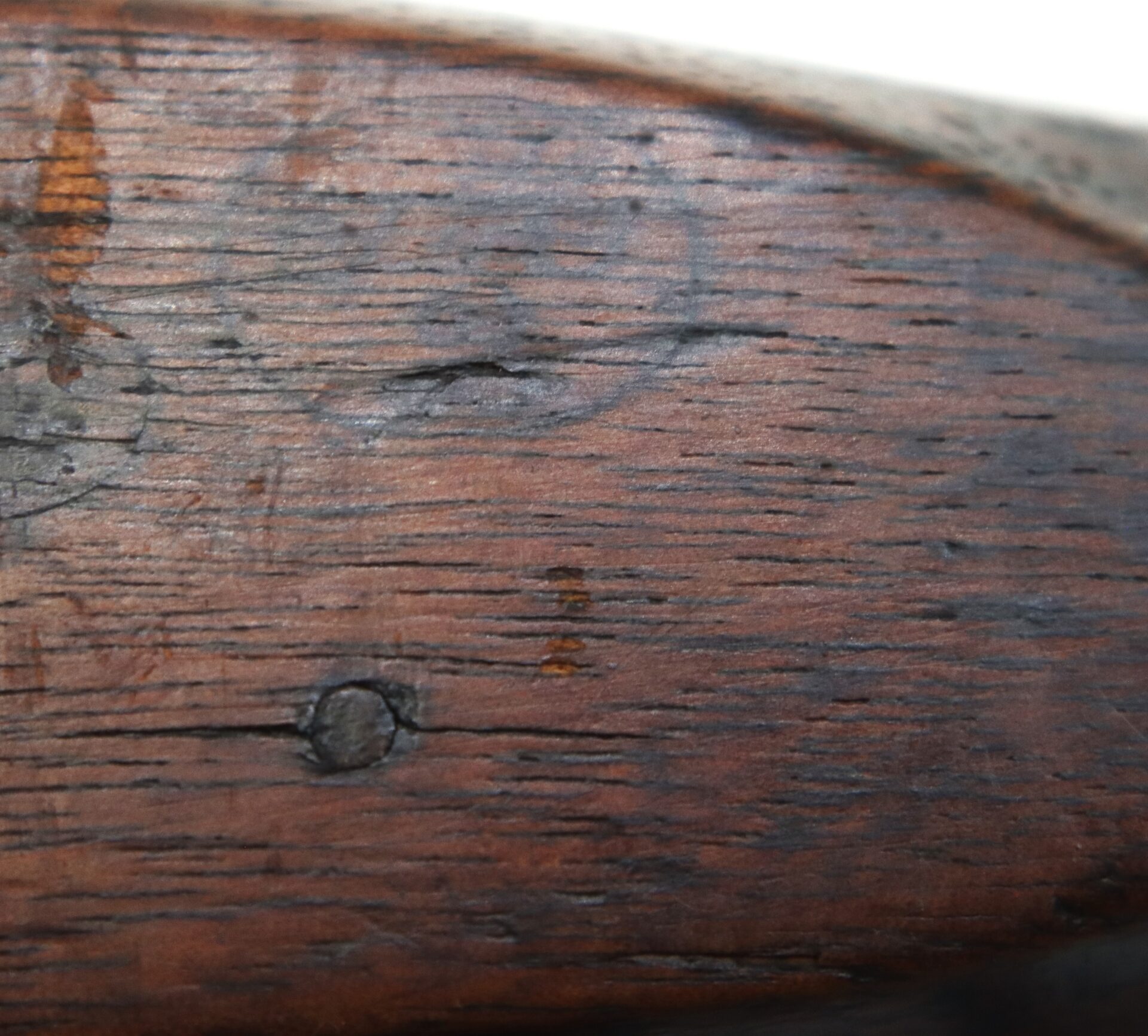 Close-up view of a weathered wooden surface with visible grain, a small nail head, a faint black marking, and minor scratches and discoloration.