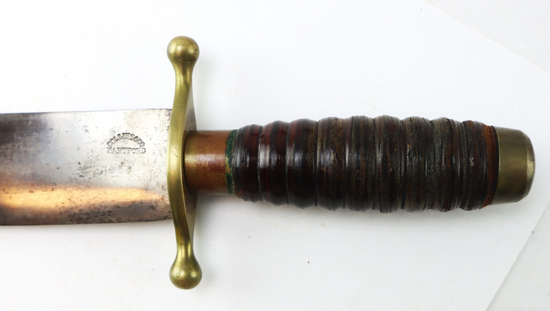 Close-up of a knife showing a worn metal blade, a brass crossguard, and a dark ribbed leather handle with brass pommel.