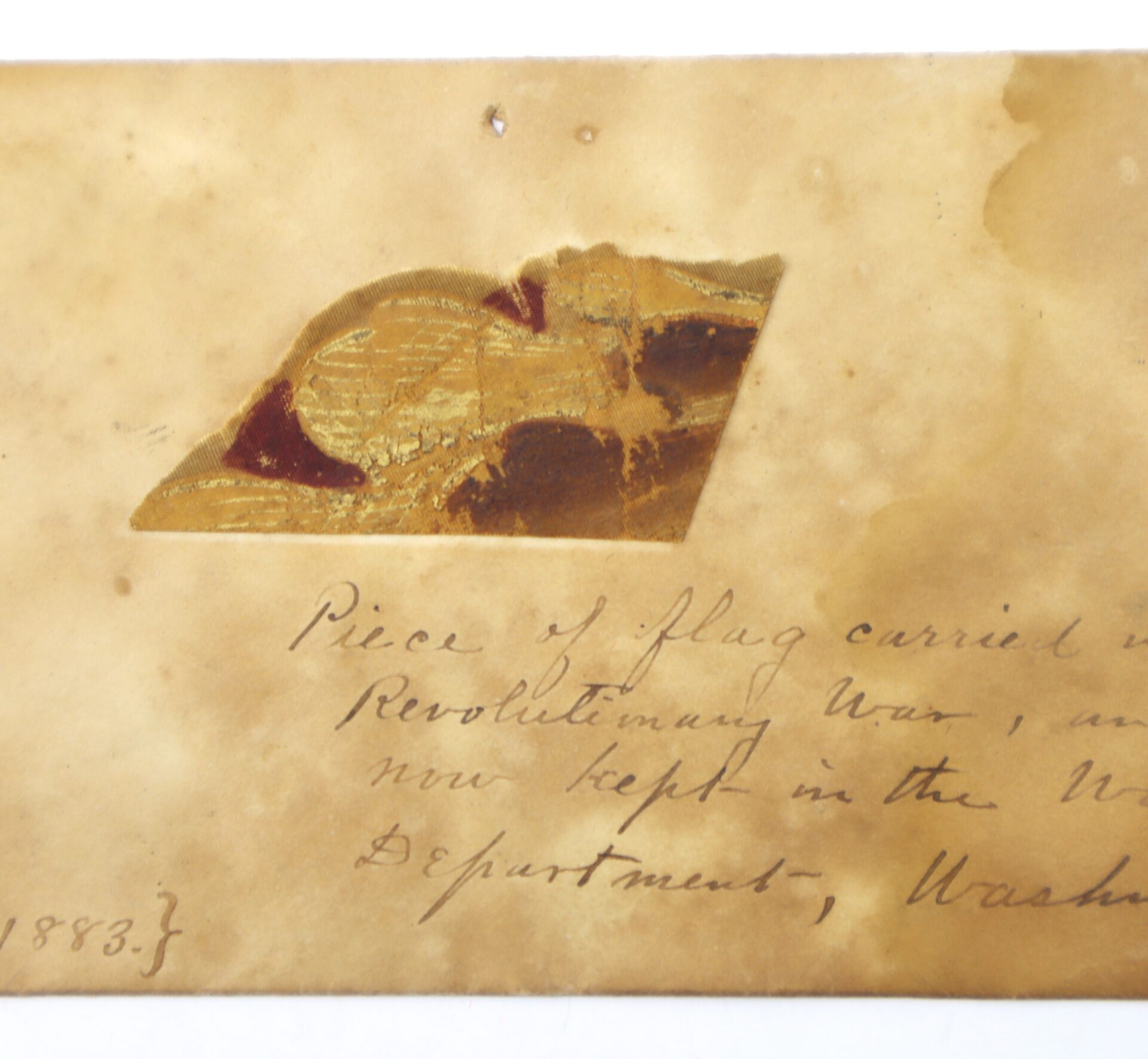 A small, worn piece of fabric is affixed to a stained, aged paper featuring cursive handwriting that identifies it as the Revolutionary War Flag Fragment with Exceptional 1883 Provenance.