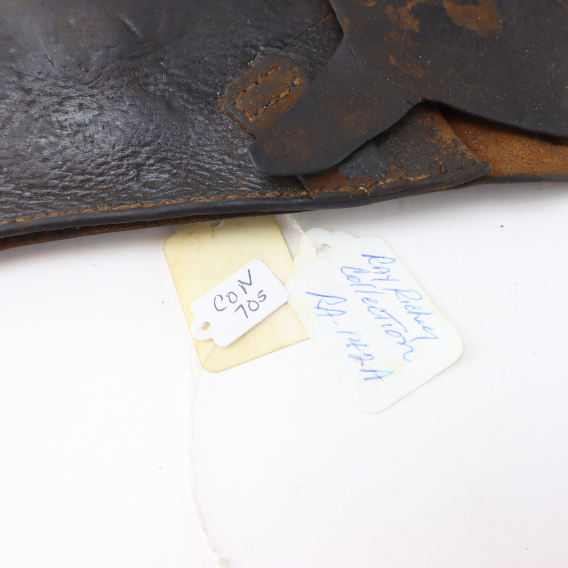 Close-up of two tags attached to a worn leather object; one tag reads “CON 705,” the other says “Pat Ritter Collection RLT 1424.”.