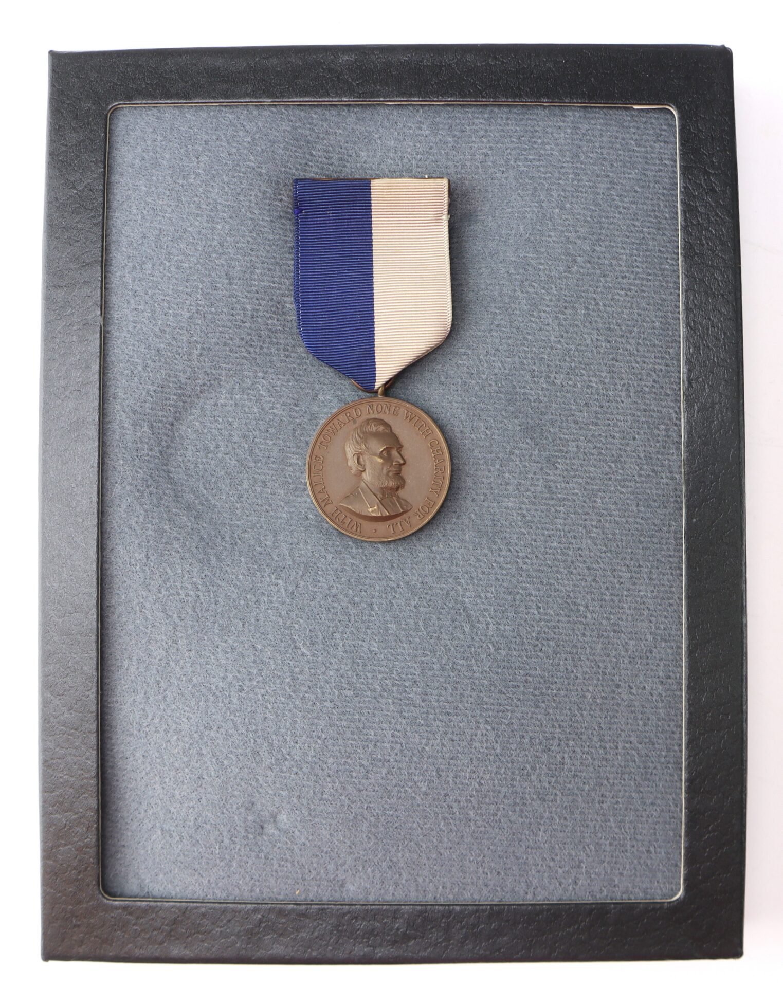 A bronze medallion with a blue and beige ribbon displayed in a black case, featuring a profile portrait and text around the edge of the medal.