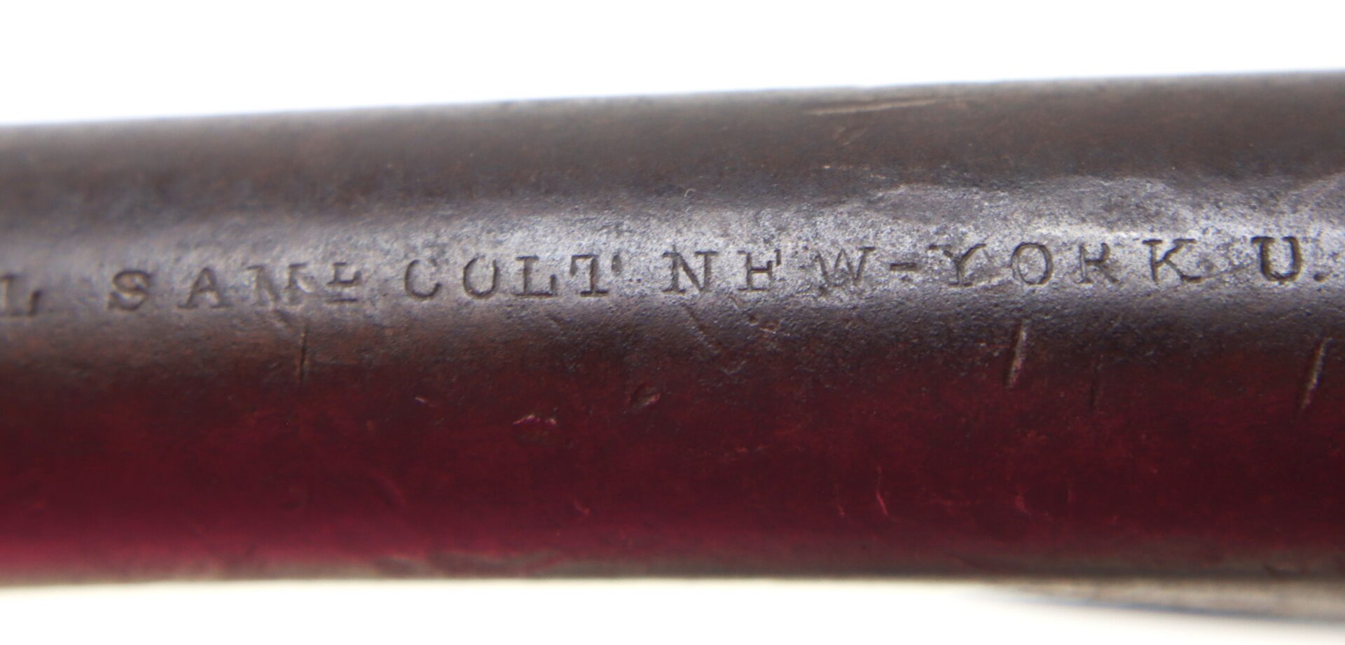 Close-up of an engraved marking on a dark metal surface reading “COLT S.A. MFG. COLT NEW-LO”. The engraving shows signs of wear and age.
