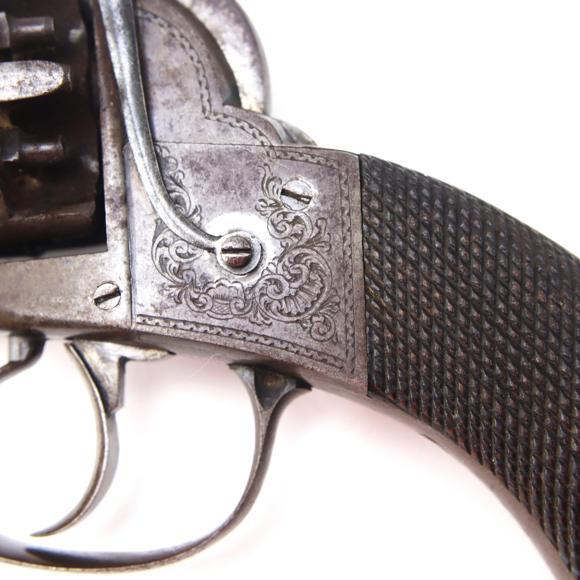 Close-up of an antique revolver showing part of the trigger, engraved metal frame, and textured dark handle against a white background.