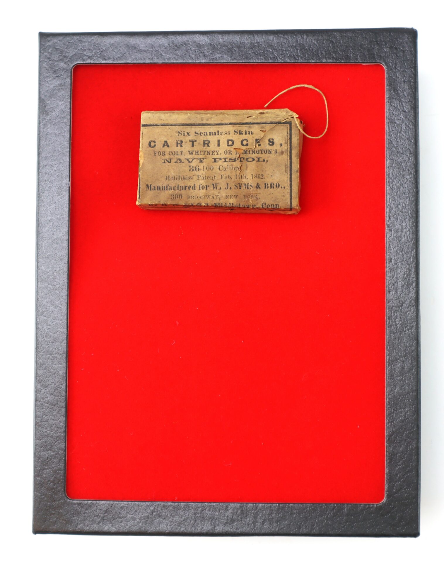 A small vintage cartridge box with a printed label is displayed against a red background within a black-edged frame.