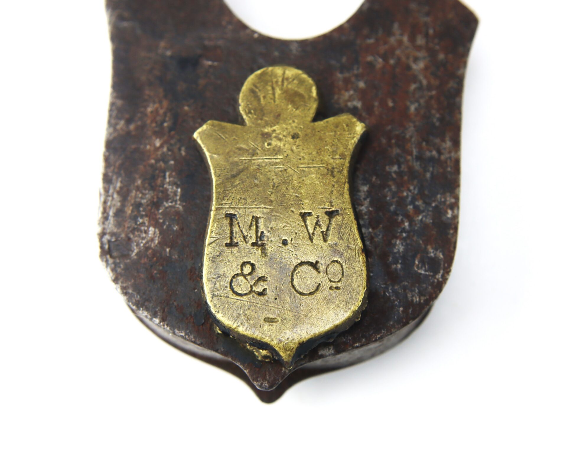 A close-up of a metal badge shaped like a shield, with the inscription “M.W & Co” engraved on its brass surface, attached to a rusted background.