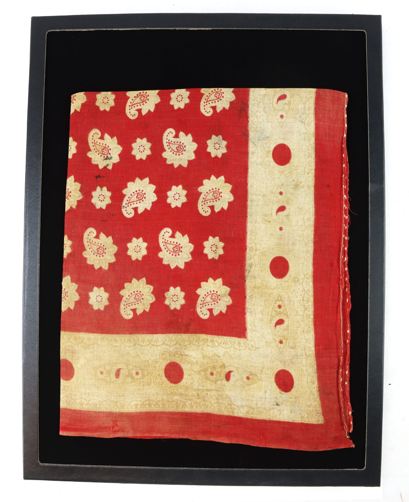 A square red cloth with a cream decorative border and paisley floral patterns, displayed flat in a black frame.