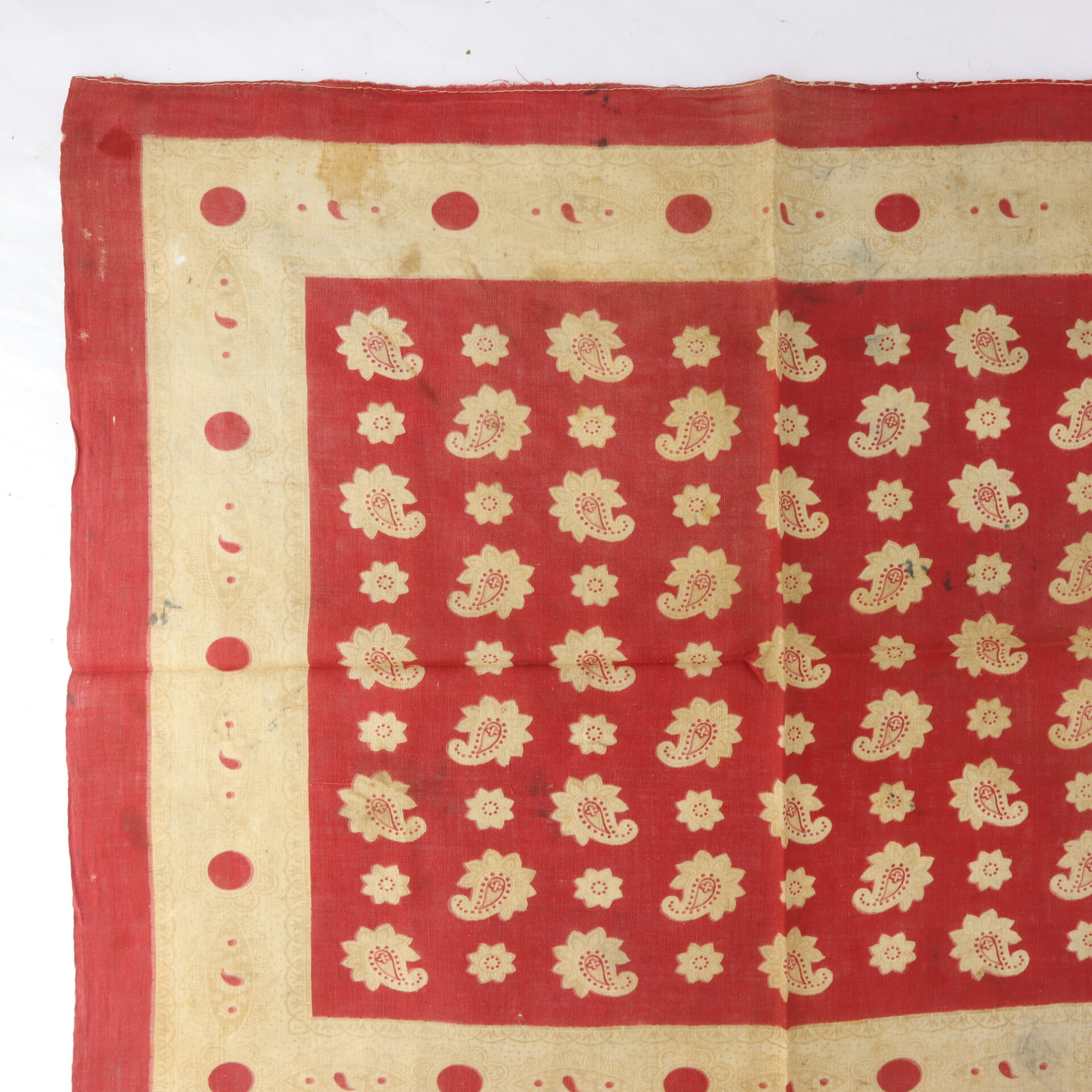 Red cloth with a beige border, decorated with a pattern of beige paisleys and flowers in the center and dots on the border. The fabric is slightly wrinkled and laid flat.