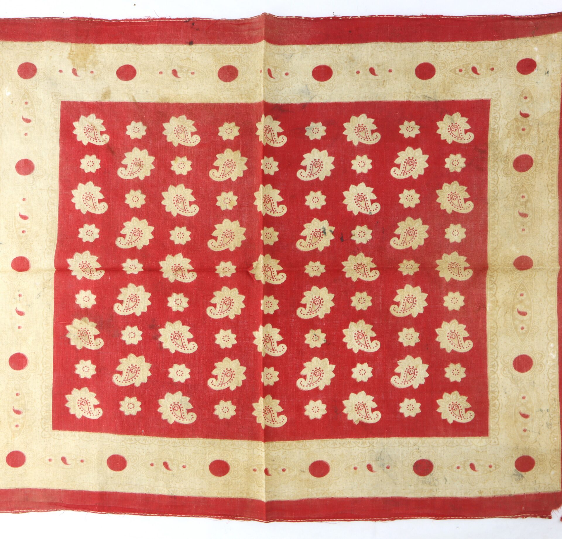 A red and beige textile with a floral and paisley pattern, featuring a beige border with red circles and a central field of repeating beige motifs on a red background.