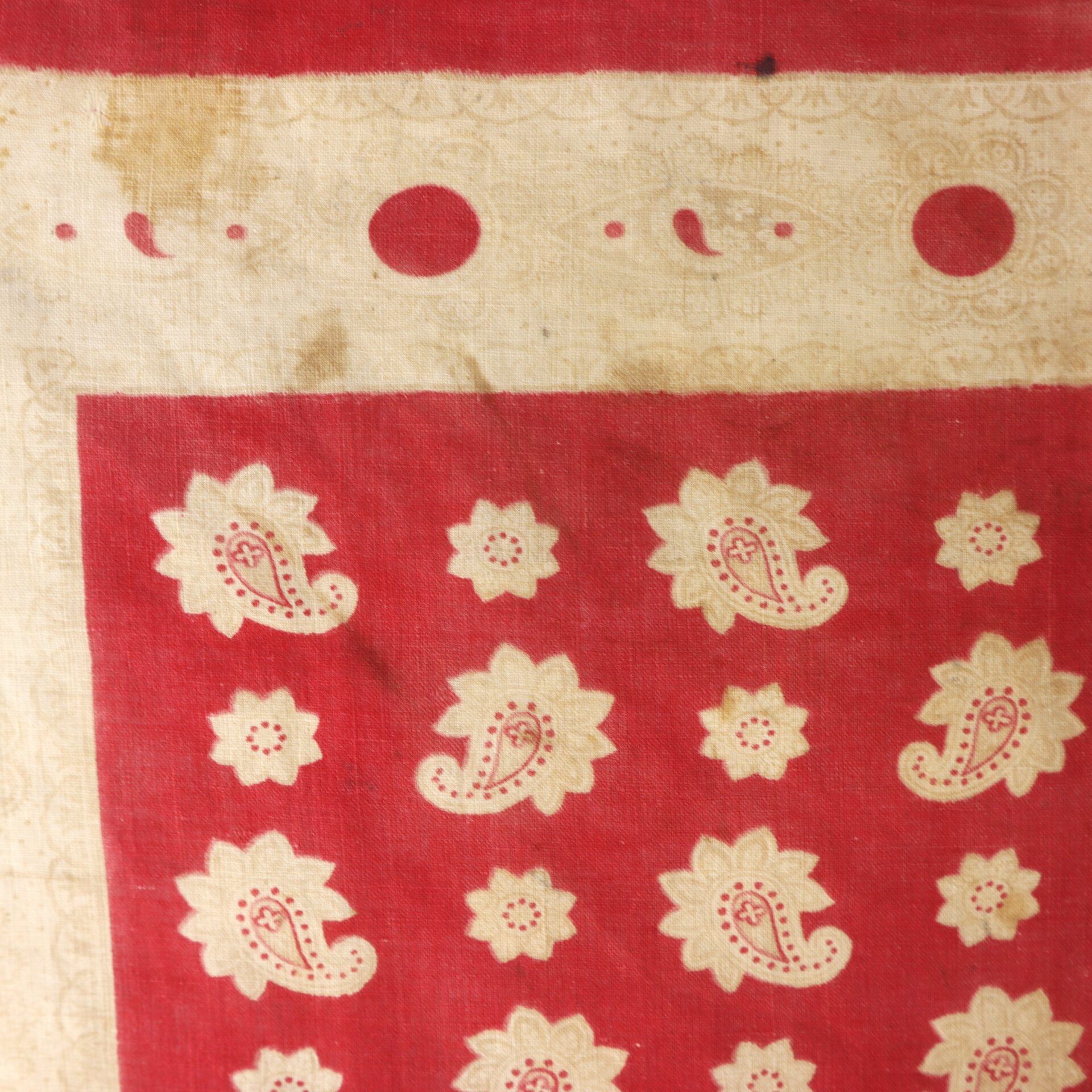 Red and beige patterned fabric with paisley and floral motifs, featuring a border with circular and paisley designs. Some visible stains and signs of wear.