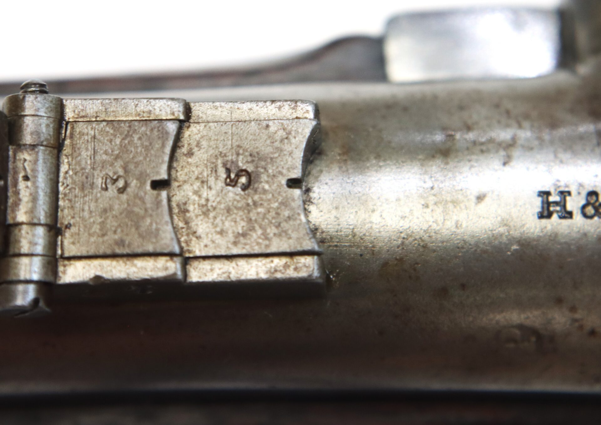 Close-up of a metal object showing numbered markings “3”, “5”, and “7” on a slotted plate, next to stamped text “H &”. The surface appears aged and slightly corroded.
