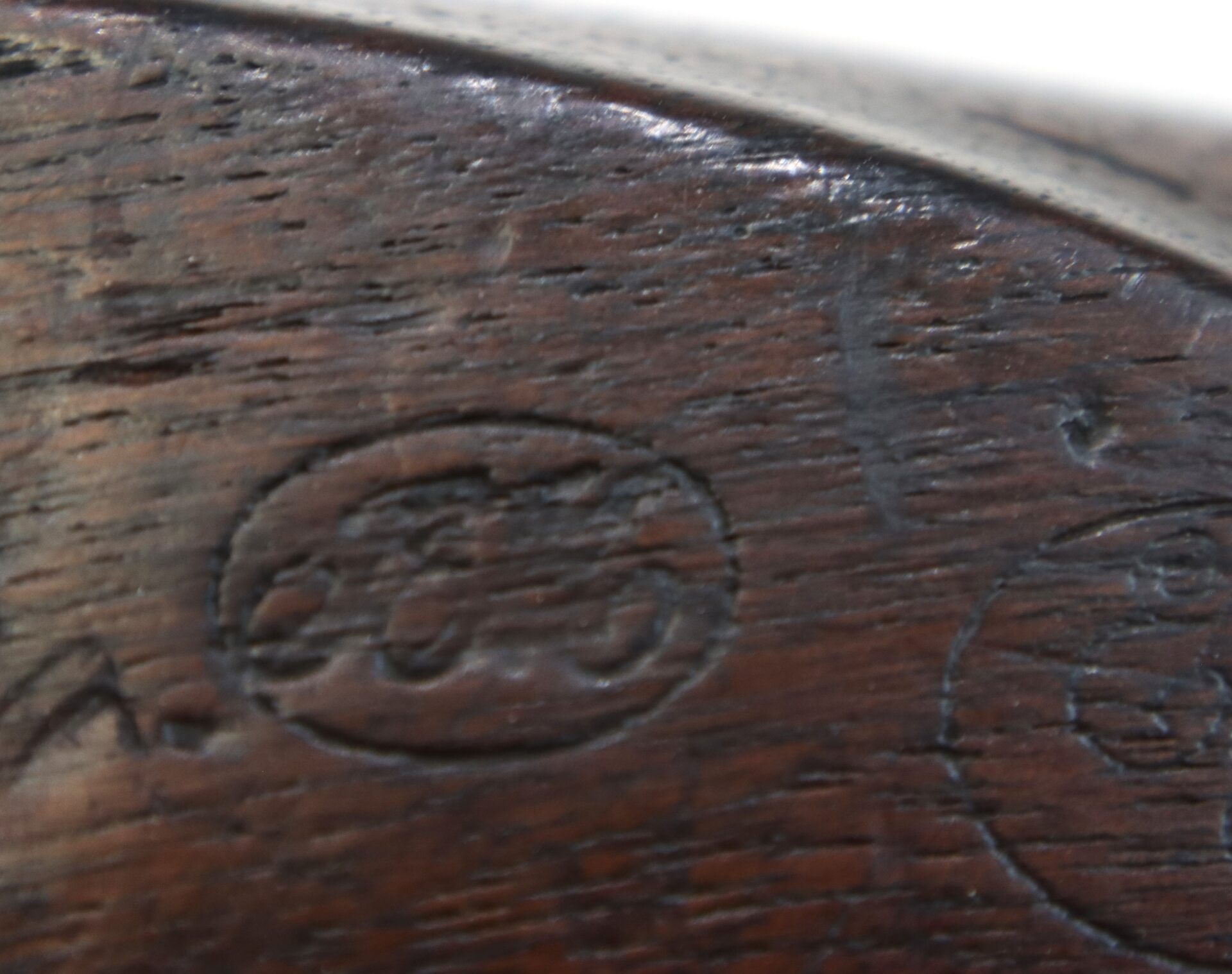 Close-up of a wooden surface with two oval-shaped stamped markings, one containing illegible script or numbers. The wood appears worn and darkened with age.