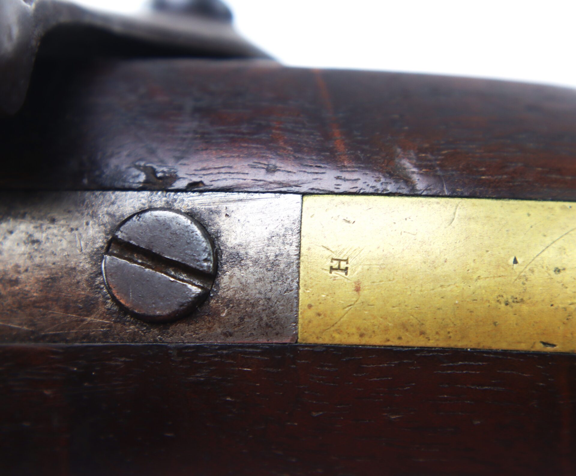 Close-up of a metal screw and a brass plate with an “H” engraving on a wooden surface, possibly part of an antique firearm.