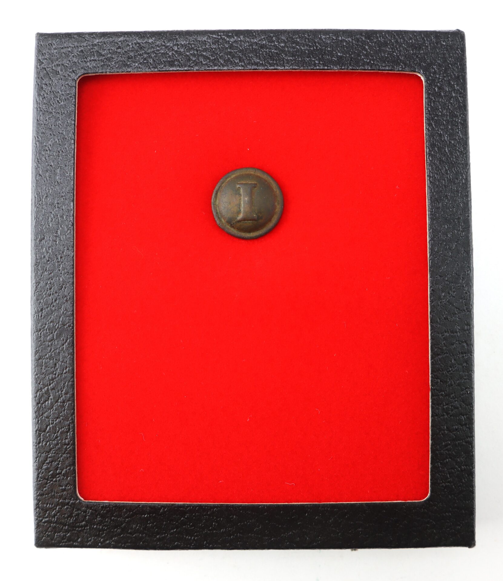 A round metal button with the letter “I” displayed in a black frame lined with red velvet.